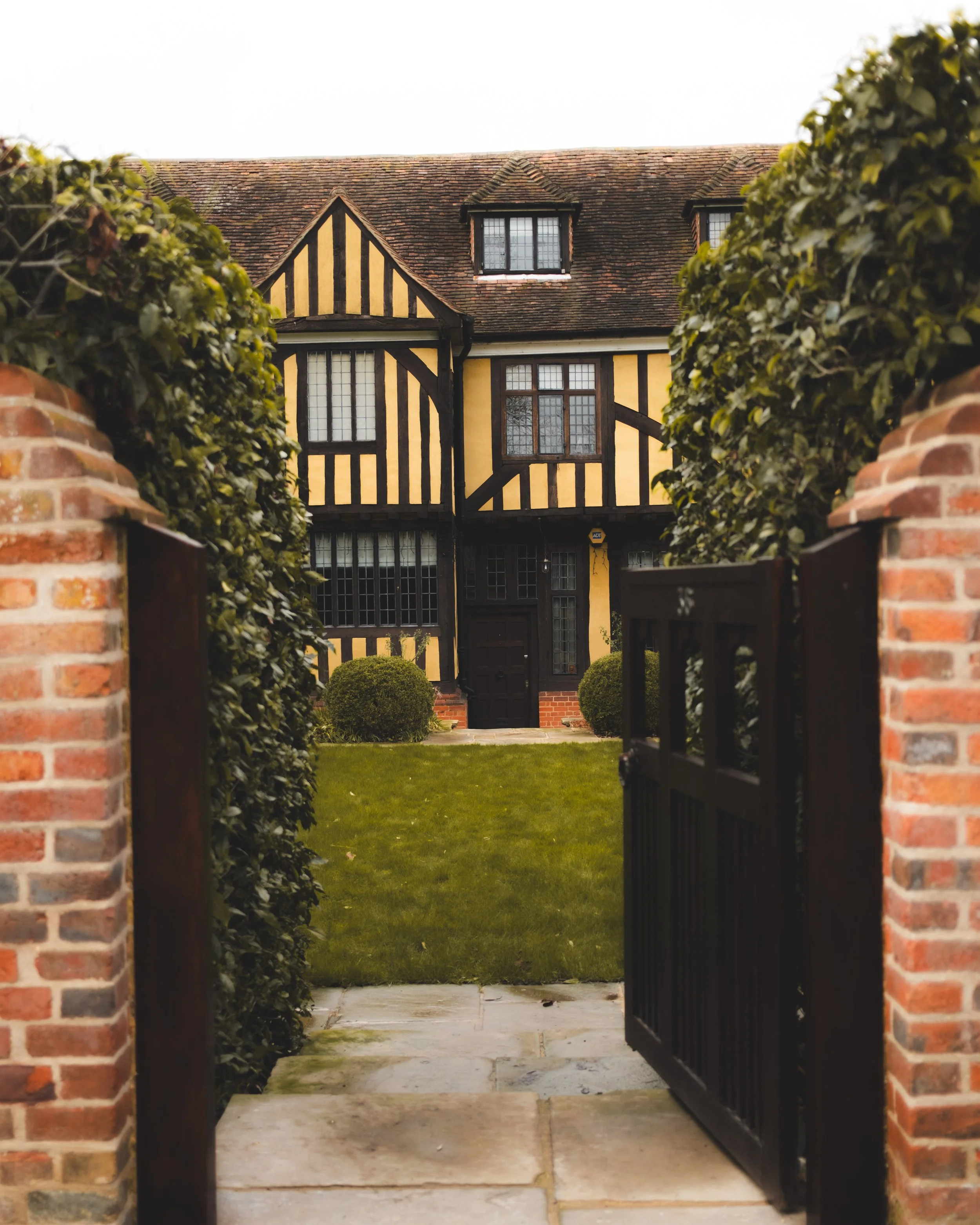 View of a house with half-timbered design, black and yellow walls, and a brick and tile roof, framed by brick pillars and a gate, with a grassy yard and bushes in front.