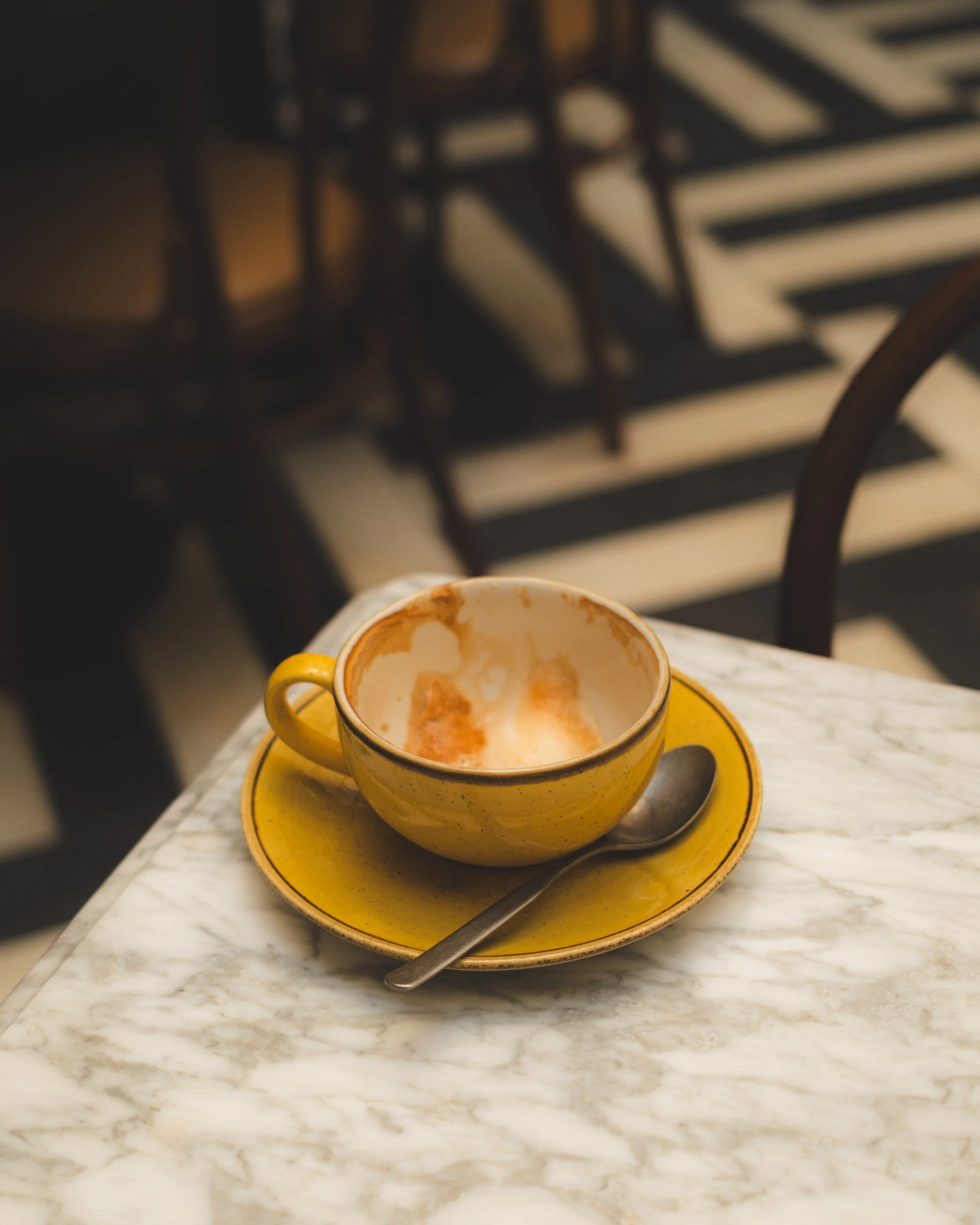 An empty yellow ceramic coffee cup with a handle, sitting on a matching yellow saucer on a white marble table. The cup has remnants of coffee or a beverage inside. A metal spoon rests on the saucer. The background features black and white striped flooring and some dark wooden chairs, slightly out of focus.
