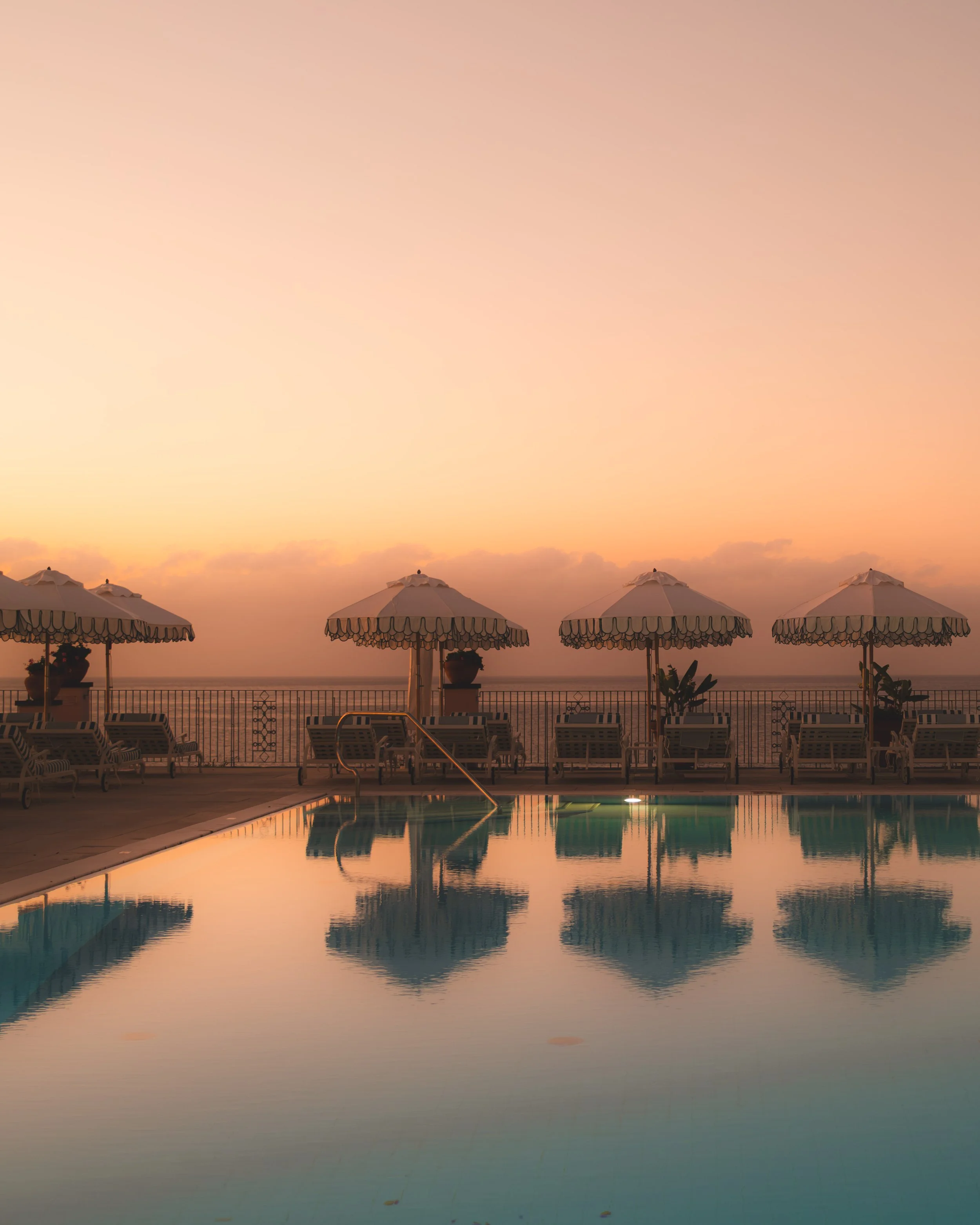 Beachside area with a pool, lounge chairs, and umbrellas during sunset. The sky has shades of pink and orange with calm ocean in the background.