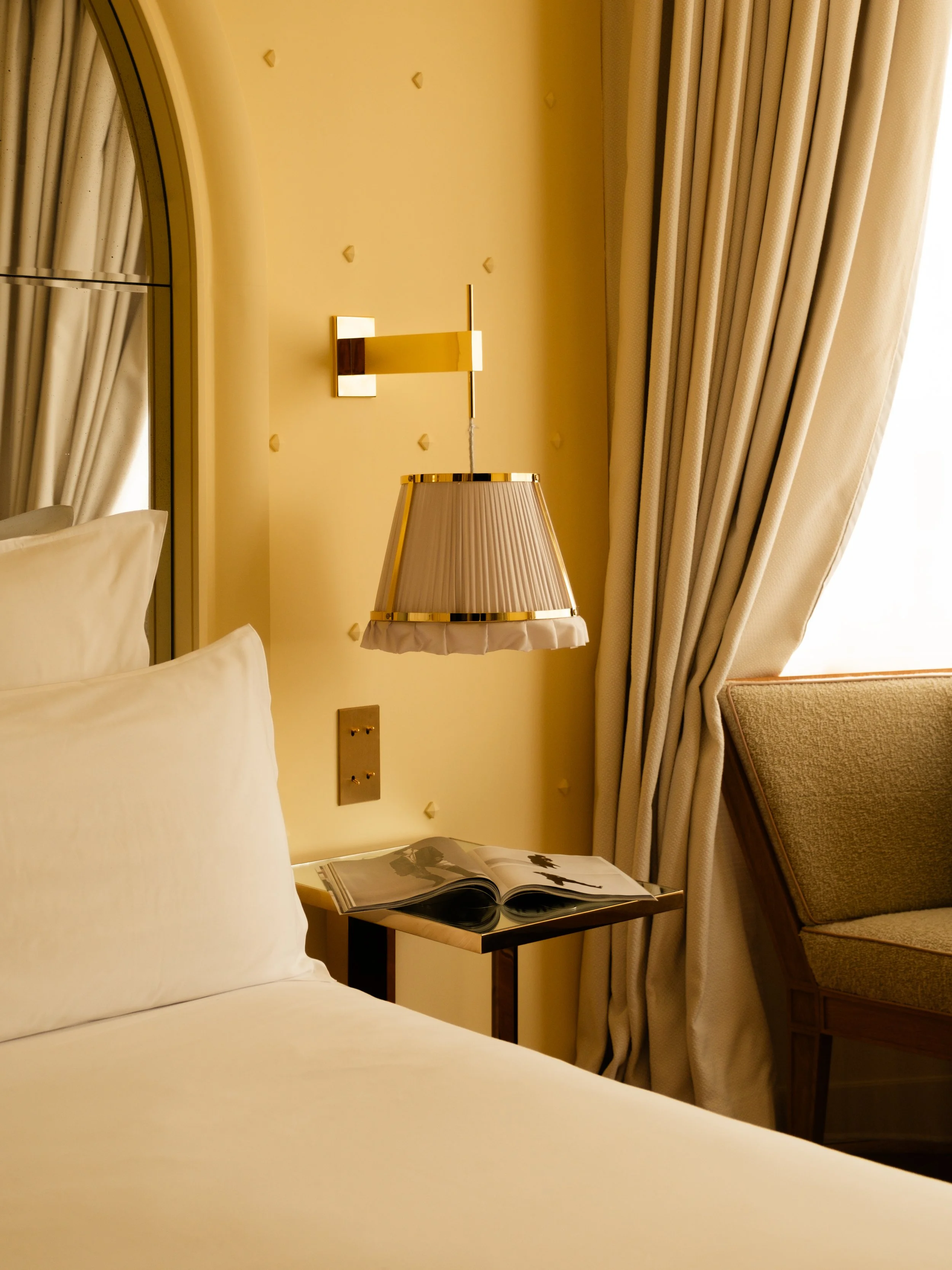 Close-up of a bed in a hotel room with a nightstand, lamp, and open magazine, beige curtains, and a window to the right. Lionel Chu - Hotel & Travel Photography