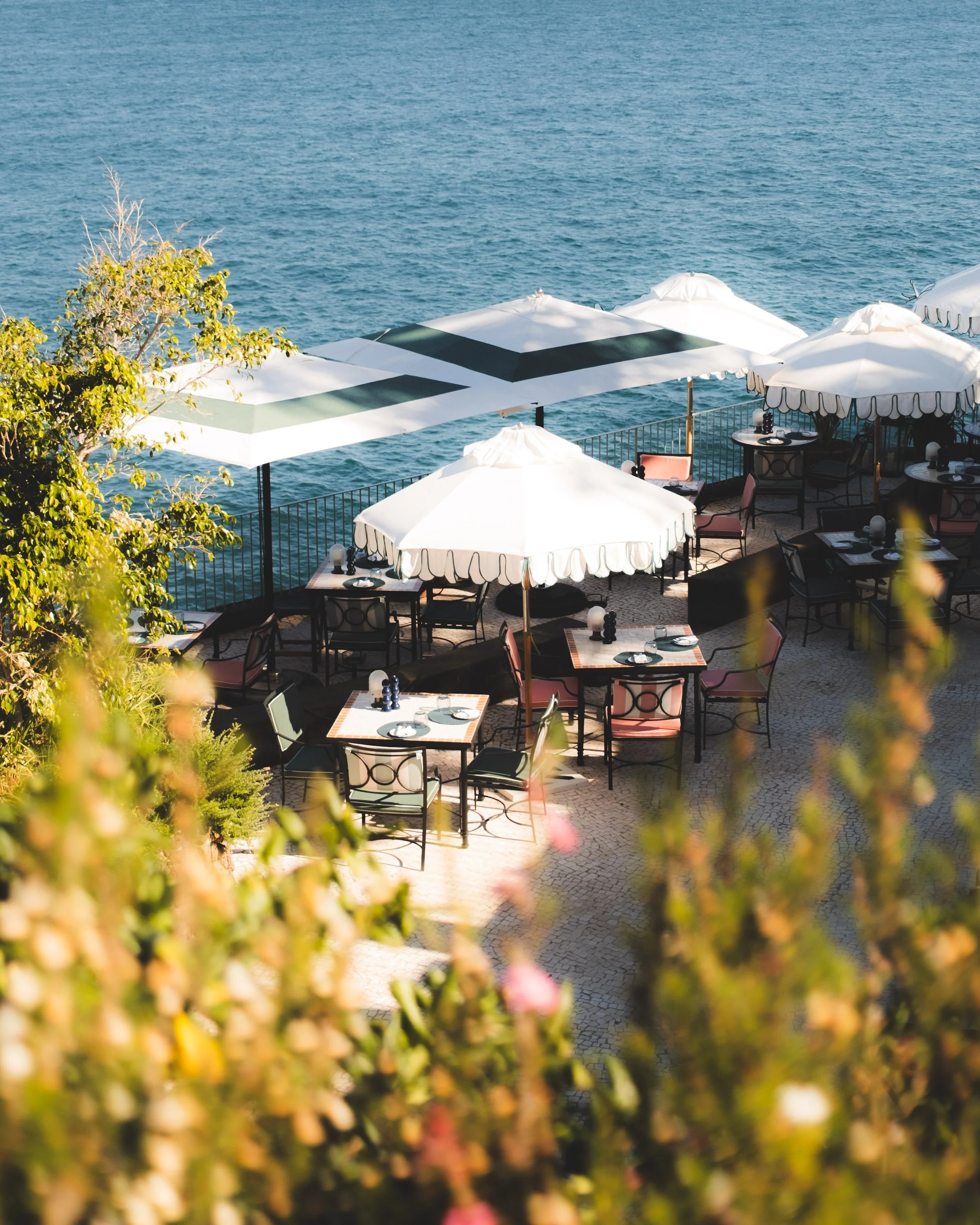 Outdoor restaurant seating area with white umbrellas and tables with dishes, overlooking a large body of water, framed by blurred trees in the foreground.