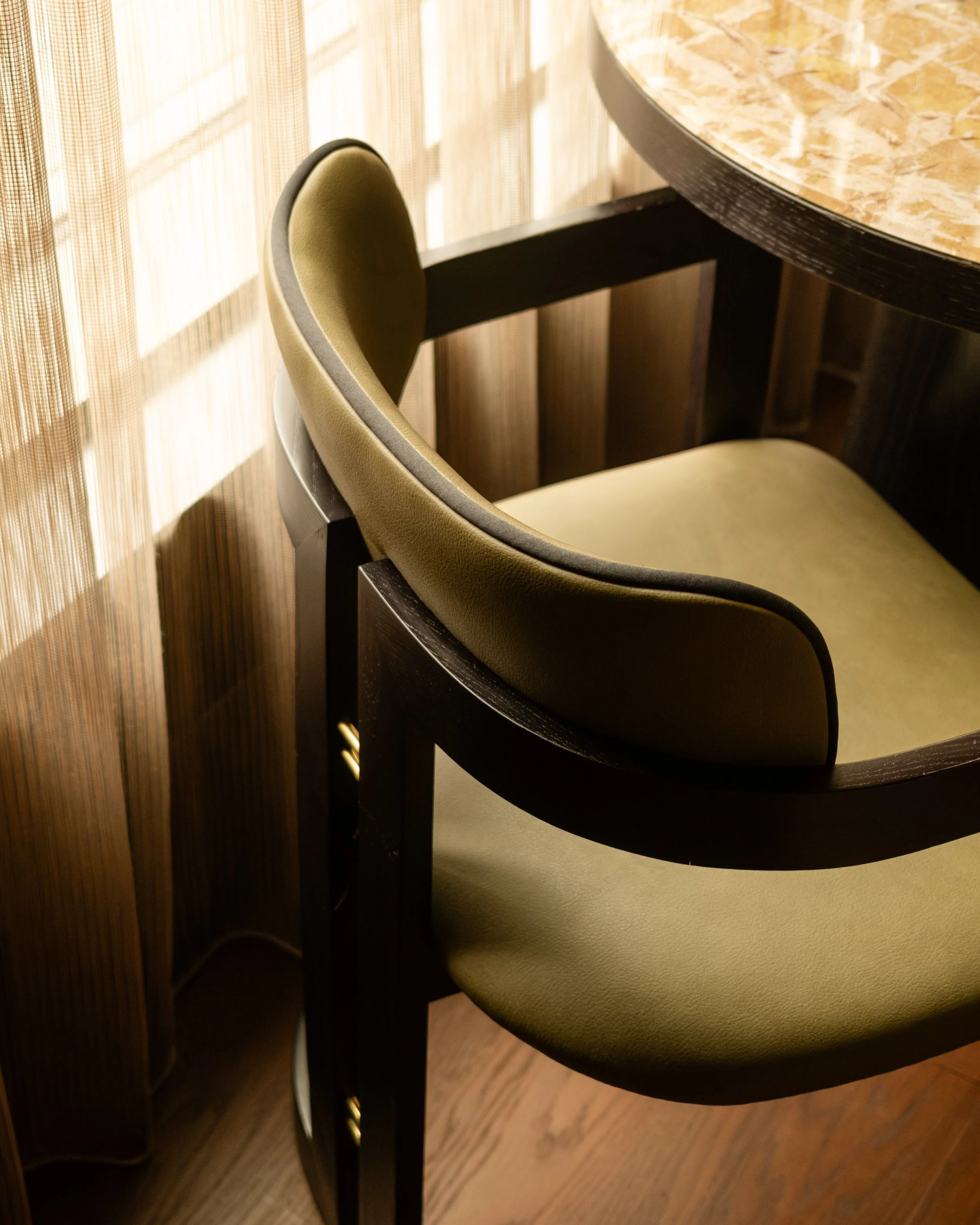 Close-up of a dining chair with green cushion and black wooden frame, next to a round marble dining table, in a room with wooden paneling and bamboo blinds. Lionel Chu - Hotel & Travel Photographer