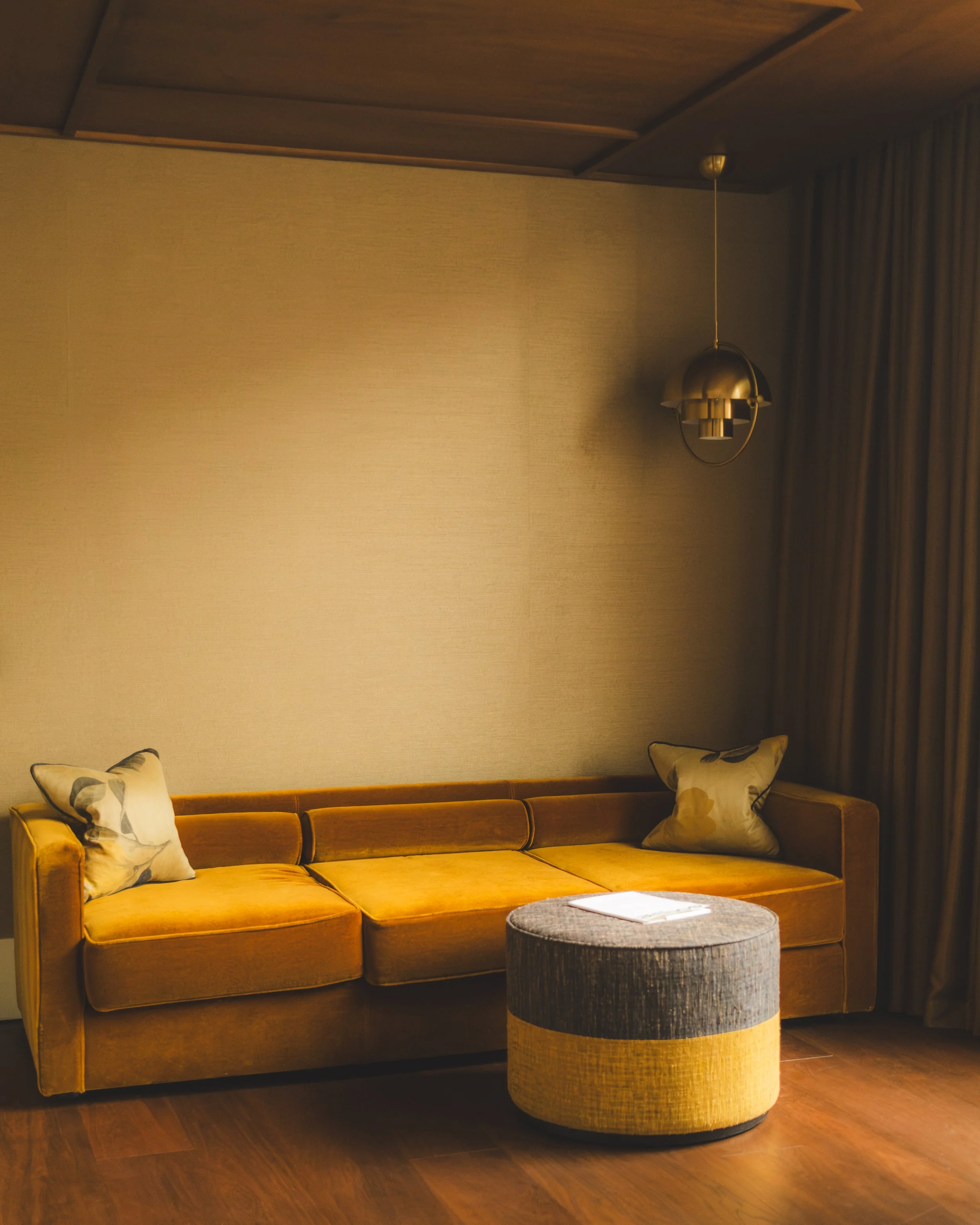 A living room with a yellow velvet sofa, patterned pillows, a round ottoman, a wall-mounted ceiling light, and curtains on the side.
