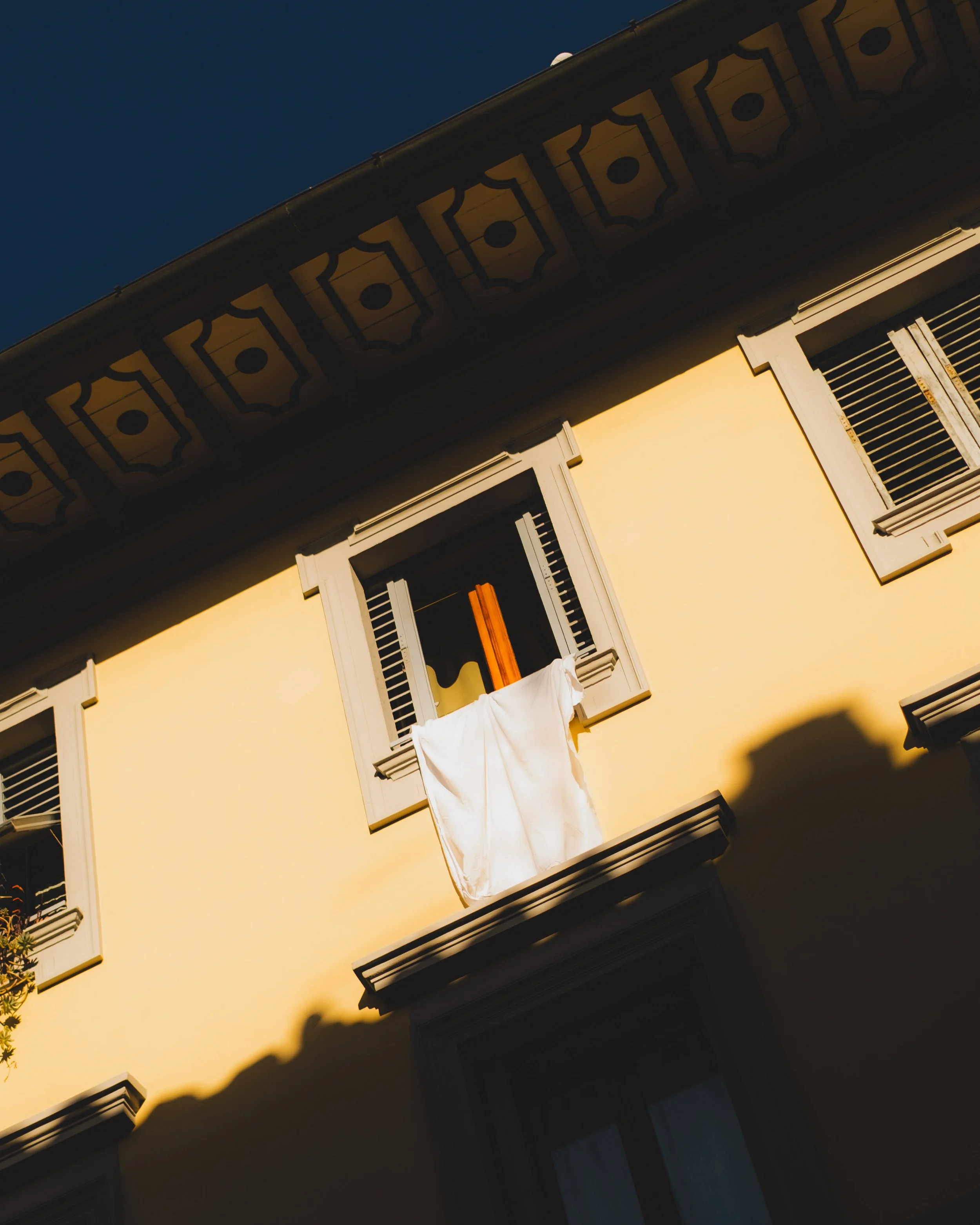 A yellow building with an open window and gray shutters, a white cloth hanging from the window, and decorative trim on the roof. Lionel Chu - Hotel & Travel Photographer