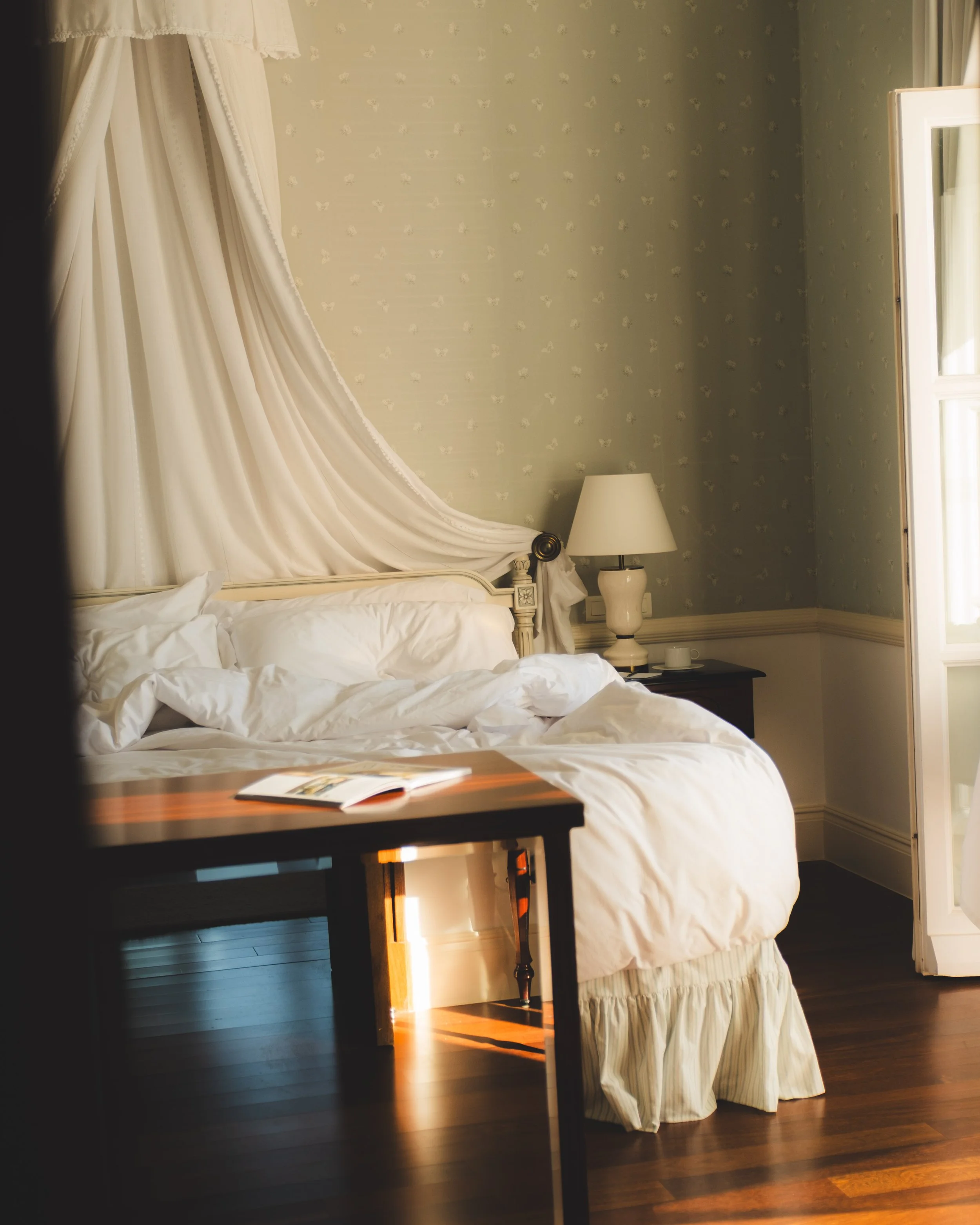 A bedroom with a bed featuring white bedding and a sheer, cream-colored canopy draped over the headboard, a small side table with a white lamp, dark wooden flooring, and a partially open window with white curtains, bathed in natural light.