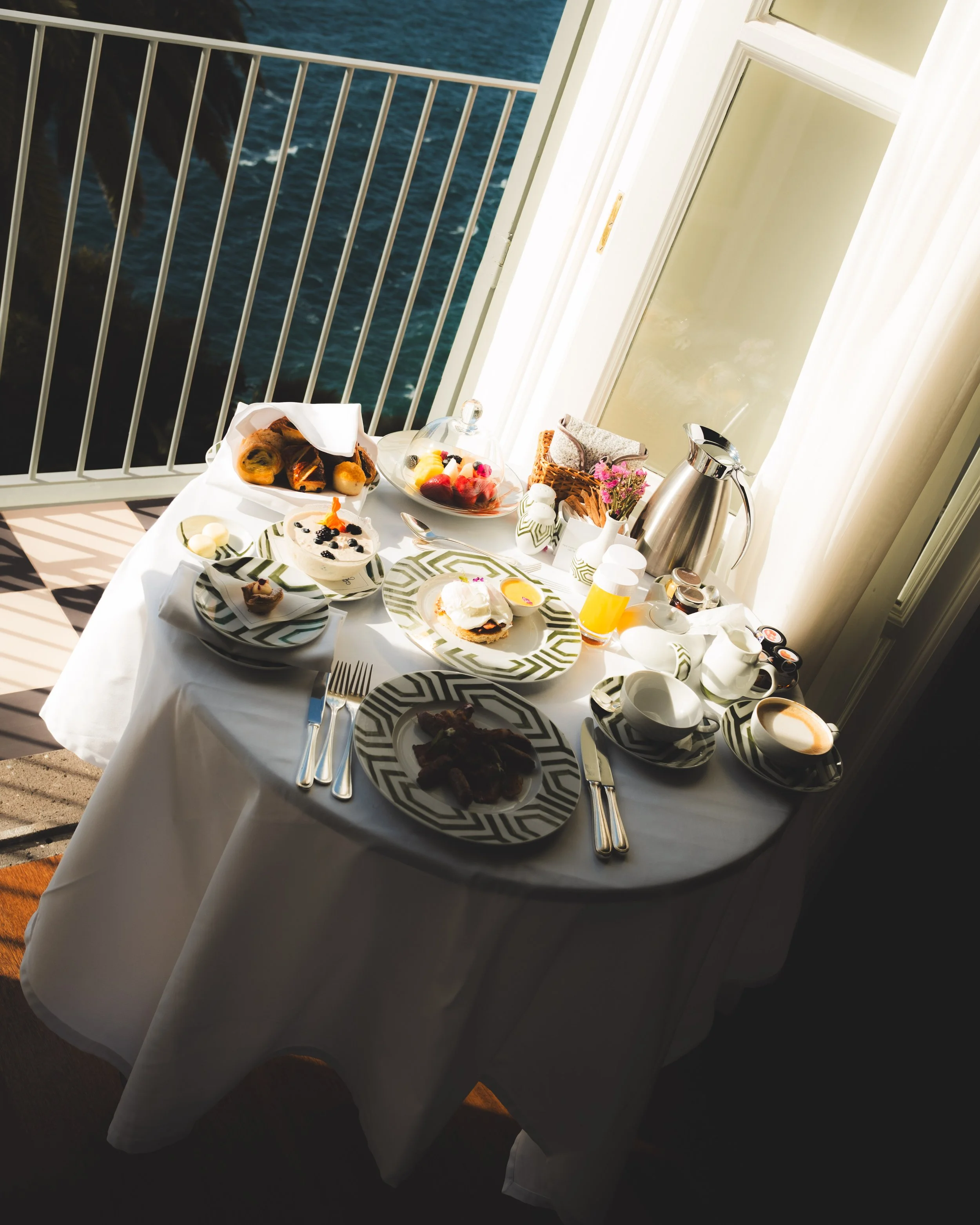A breakfast table set with plates of food, cups, a teapot, and a glass of orange juice on a balcony overlooking the ocean, with a white railing and sunlight streaming in.