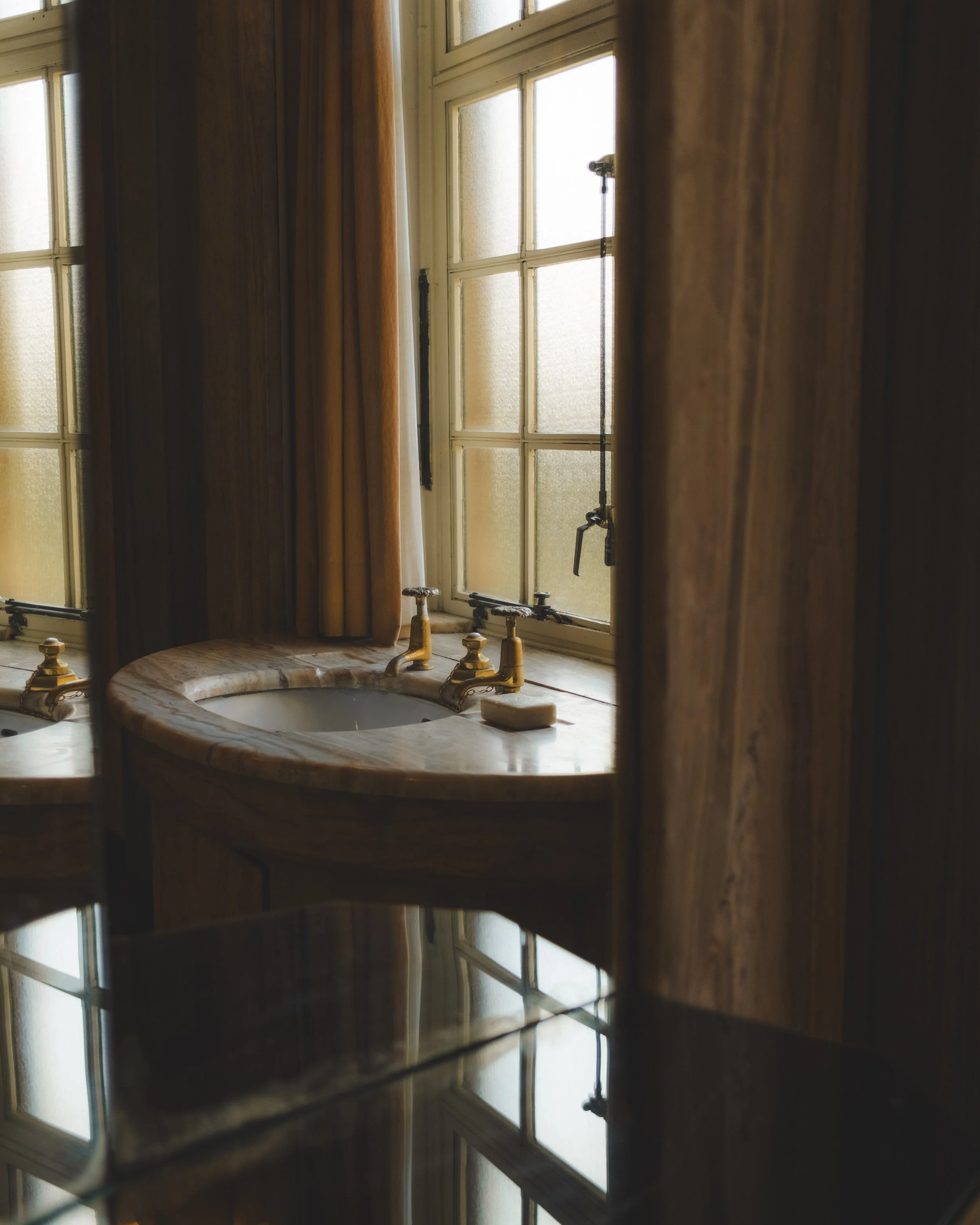 Close-up of a bathroom or kitchen counter with a sink, gold-colored faucets, and a bar of soap, with a window in the background.
