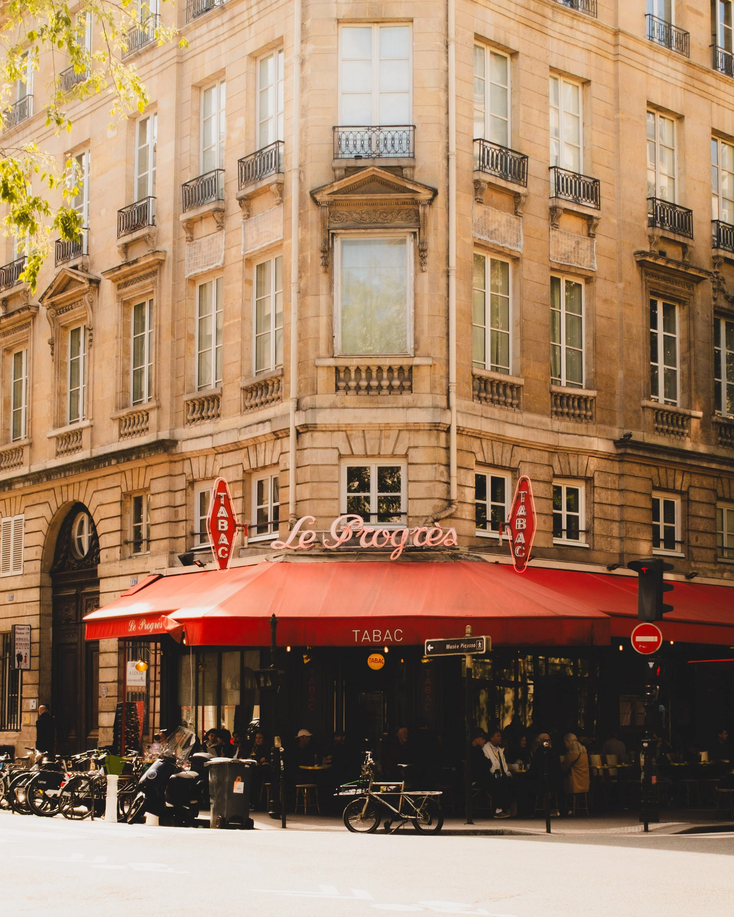 A busy Parisian cafe named Le Progrès located on a street corner with a red awning and outdoor seating, surrounded by bicycles and pedestrians, with a classic European building in the background. Lionel Chu - Hotel & Travel Photographer