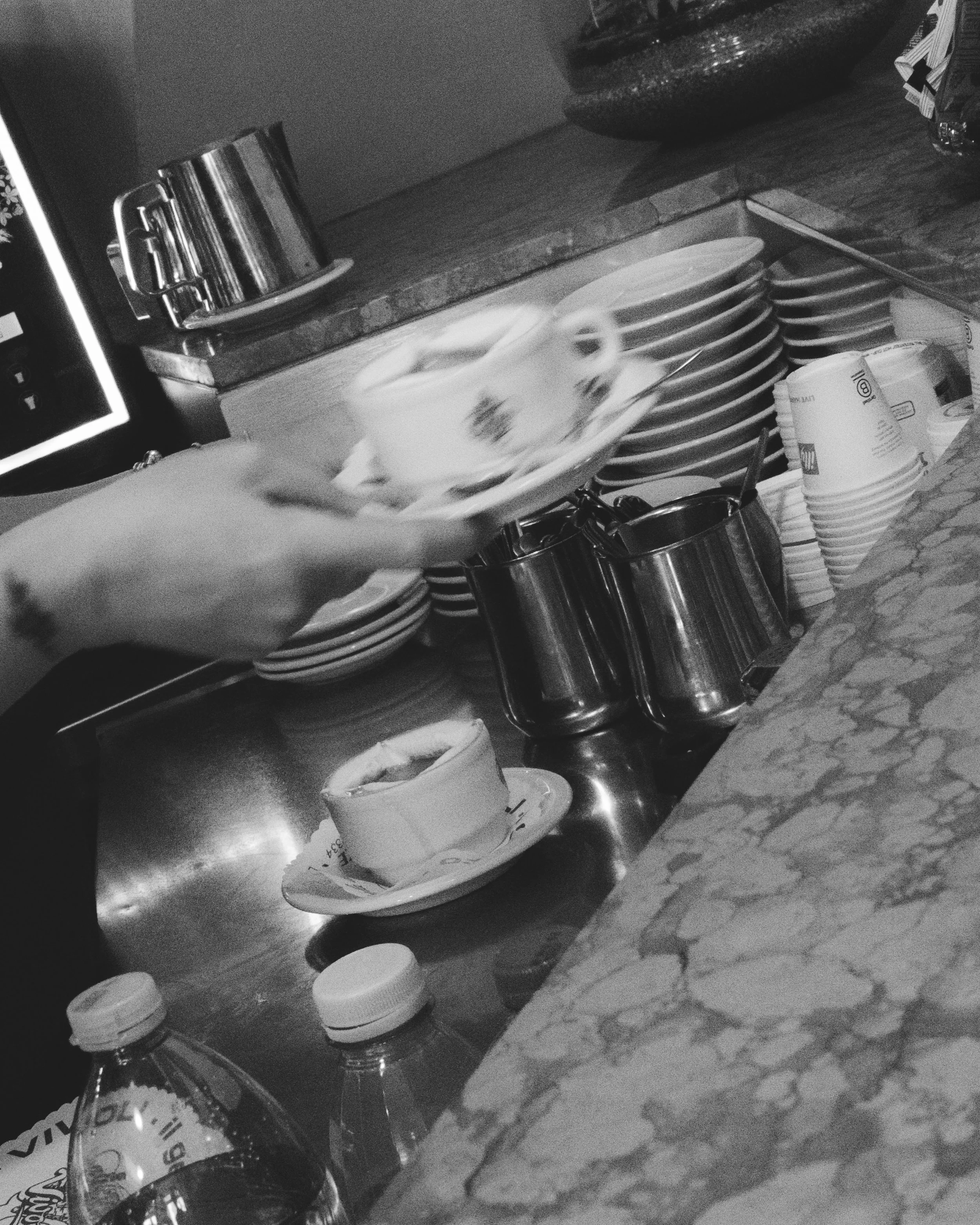 A person serving a cup of coffee or tea in a cafe with stacked plates, cups, and water bottles on a marble counter. Lionel Chu - Hotel & Travel Photographer