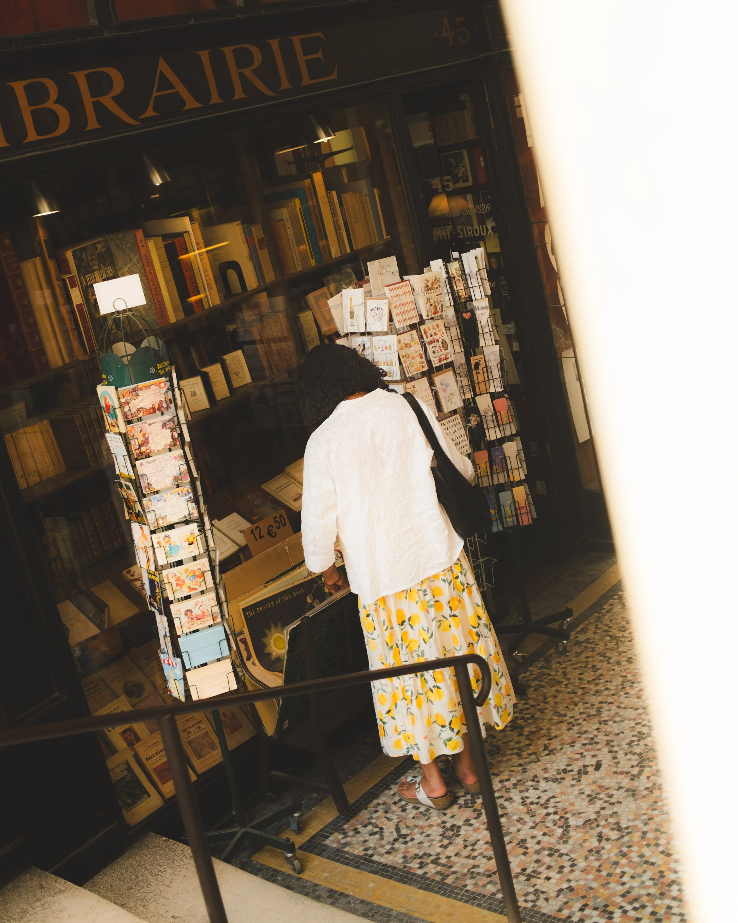 A woman wearing a white jacket and floral dress is shopping inside a bookstore. The store has shelves filled with books and a display of greeting cards. 