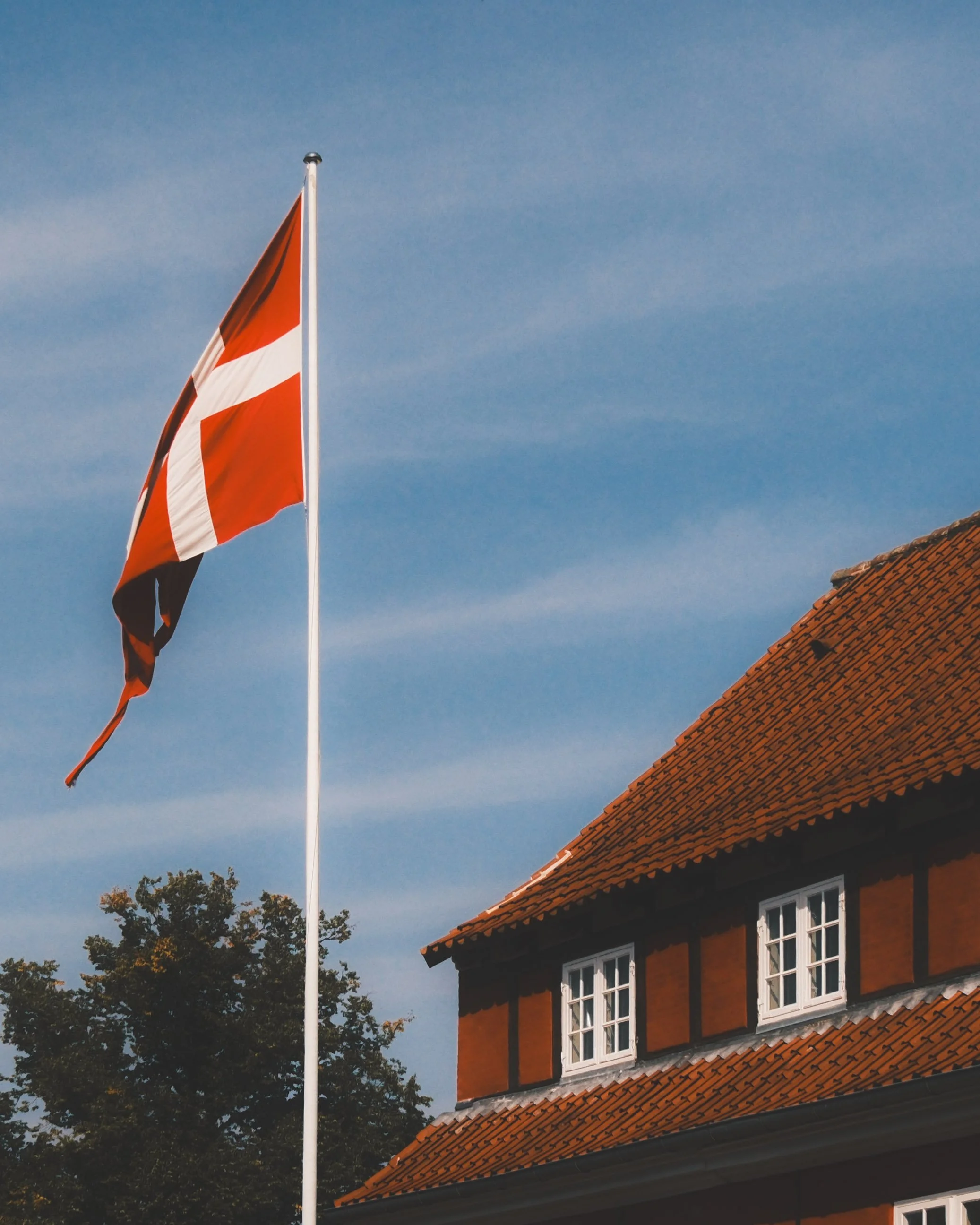 A red and white flag on a tall flagpole next to a traditional European-style house with a steep orange tiled roof and multiple white-framed windows, set against a clear blue sky.