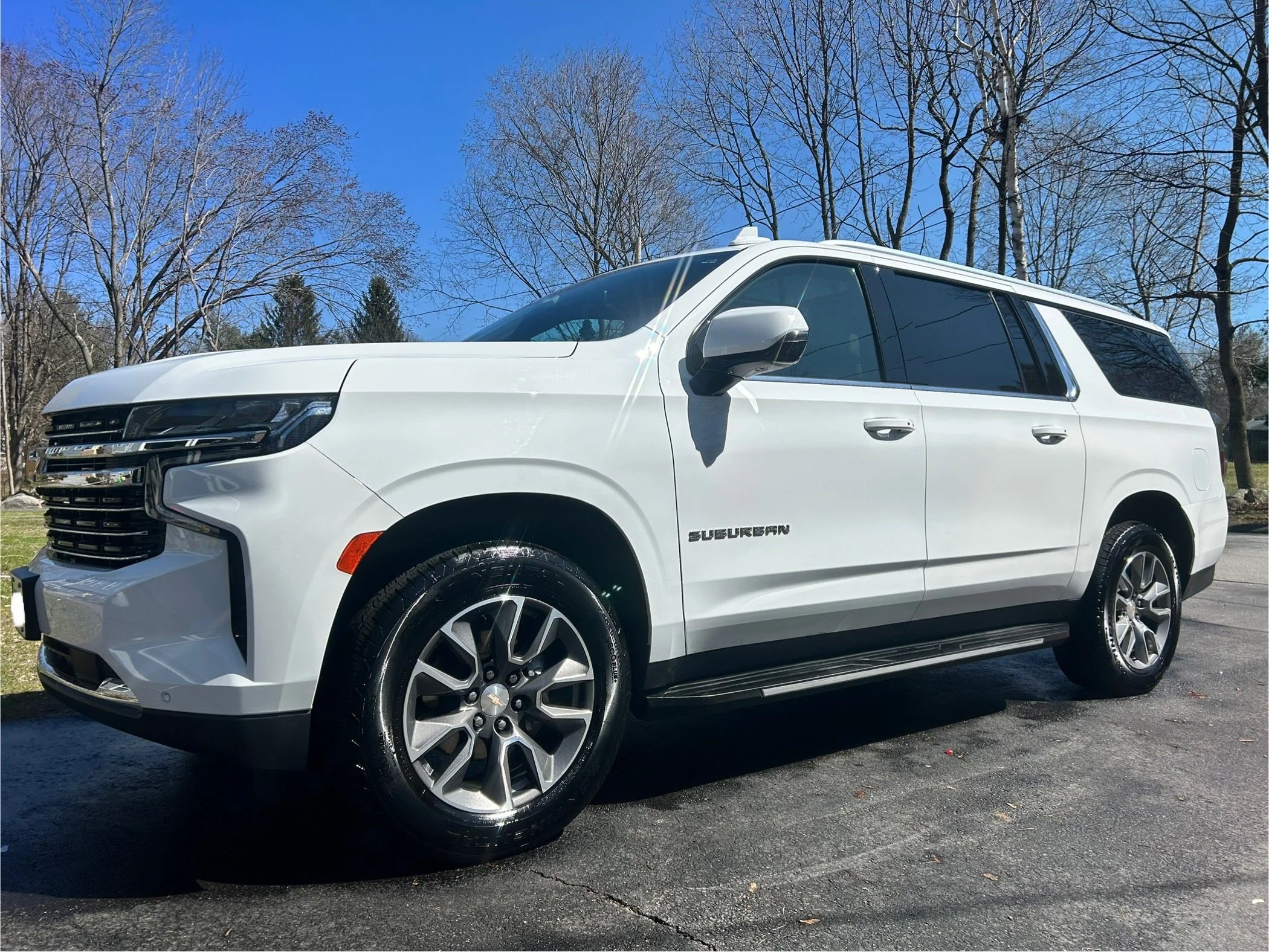 White Chevrolet Suburban SUV parked on asphalt with leafless trees and clear blue sky in the background.