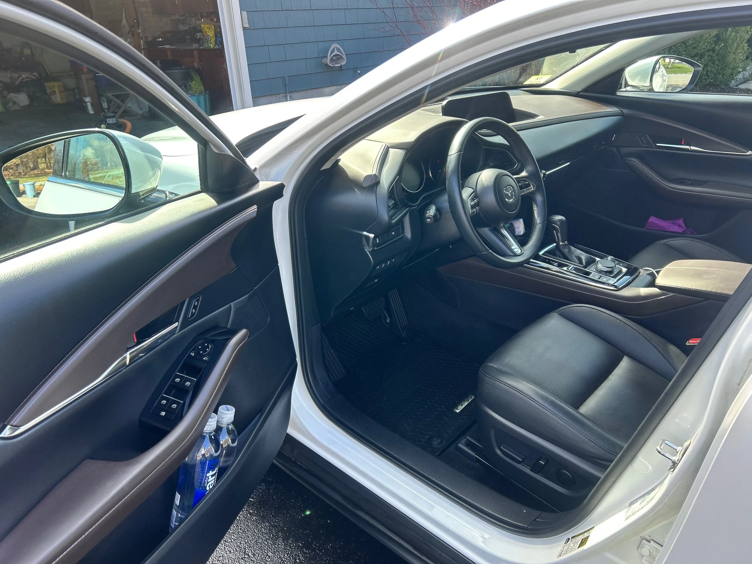 Interior of a white car, showing the driver's side with black leather seats, steering wheel, gear shift, and center console. The driver door contains window controls and two water bottles on the door pocket.