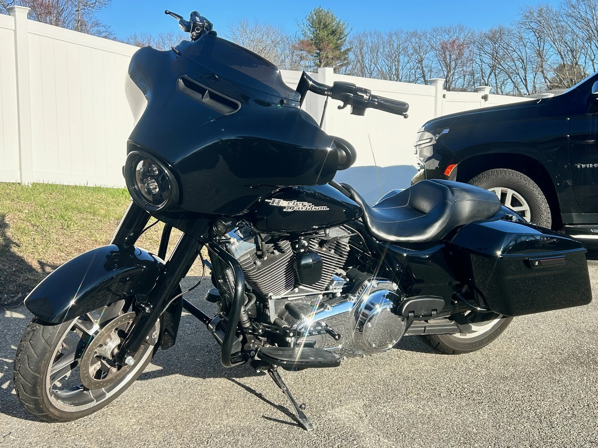 Black Harley Davidson touring motorcycle with large front fairing and saddlebags parked on a paved surface in front of a white fence and a black vehicle, with trees and a clear blue sky in the background.