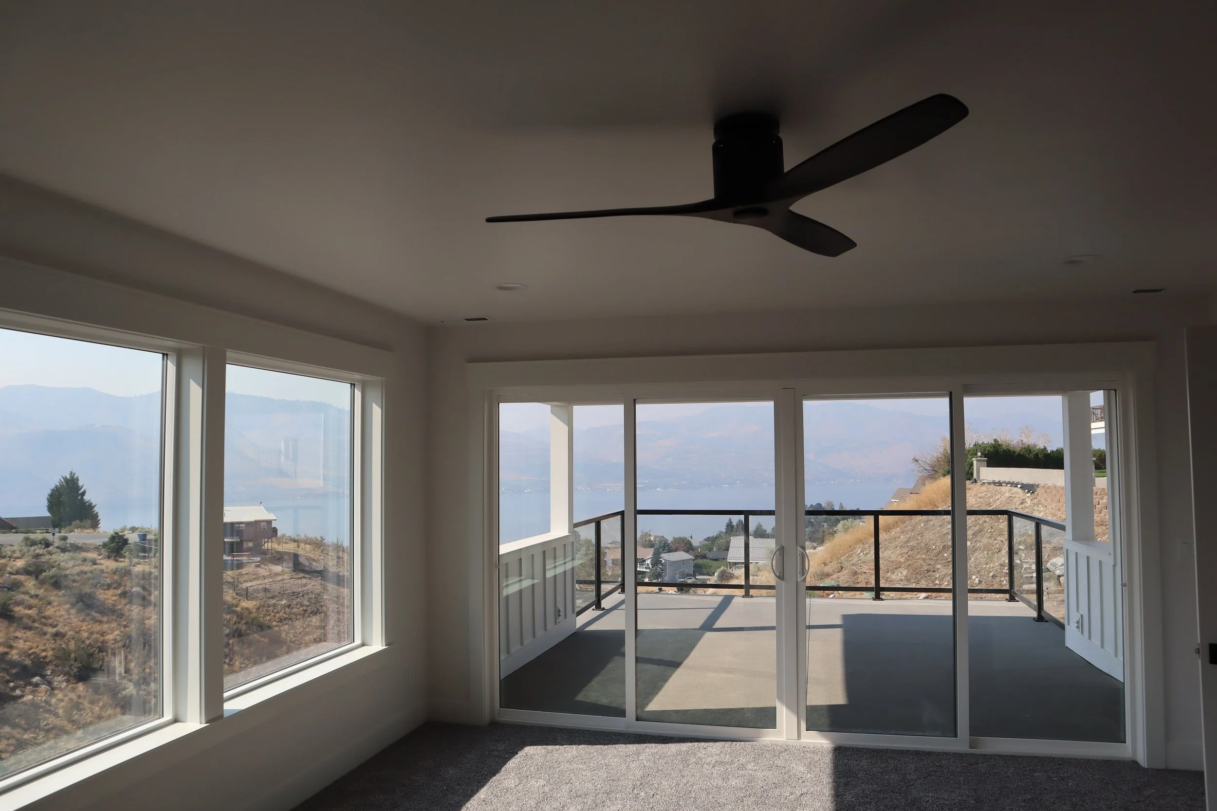 Living room with large windows and sliding glass doors overlooking a scenic outdoor view with hills and water, featuring a ceiling fan.