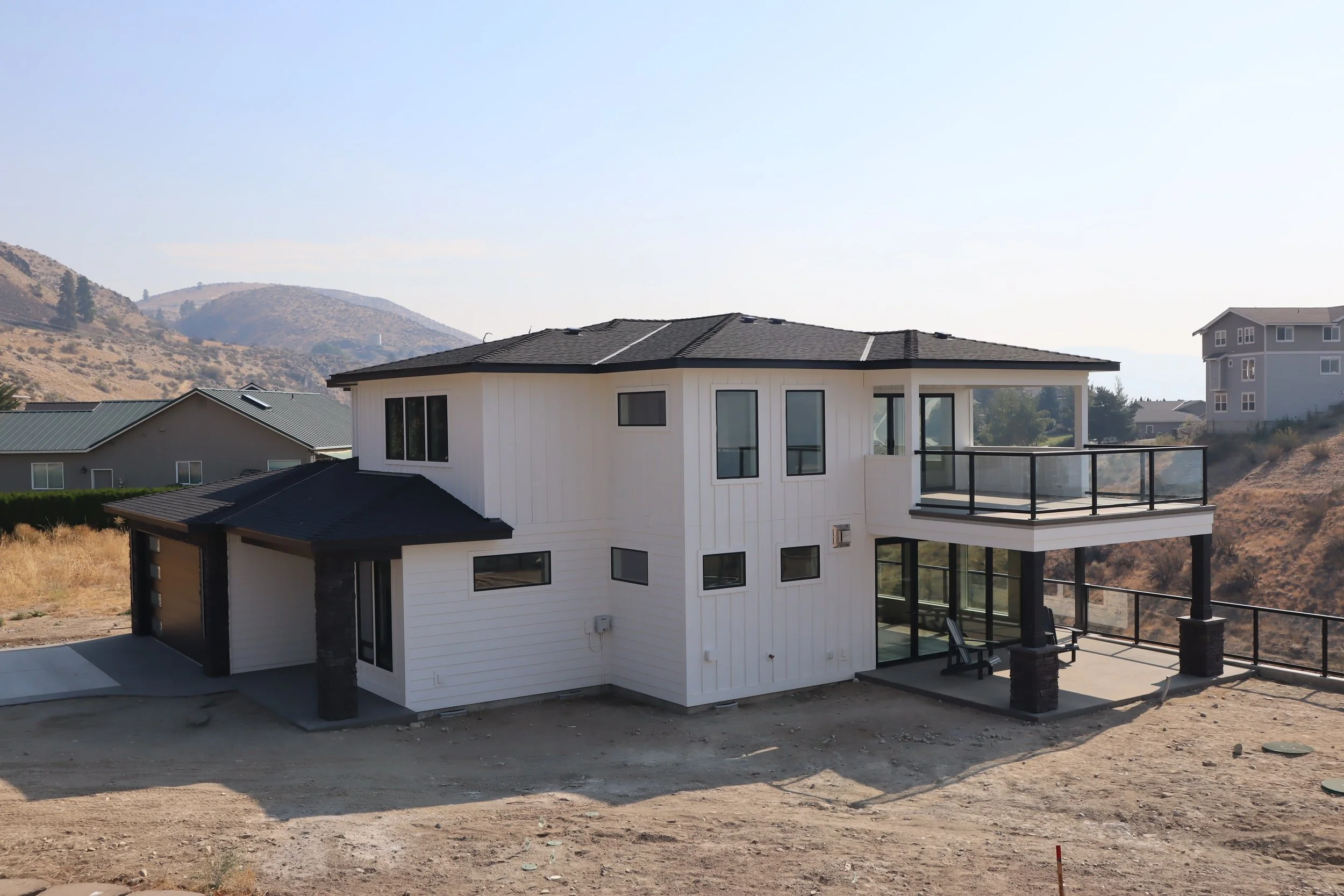 Modern white two-story house with balcony, black roof, and large windows, situated in a hilly, rural area.