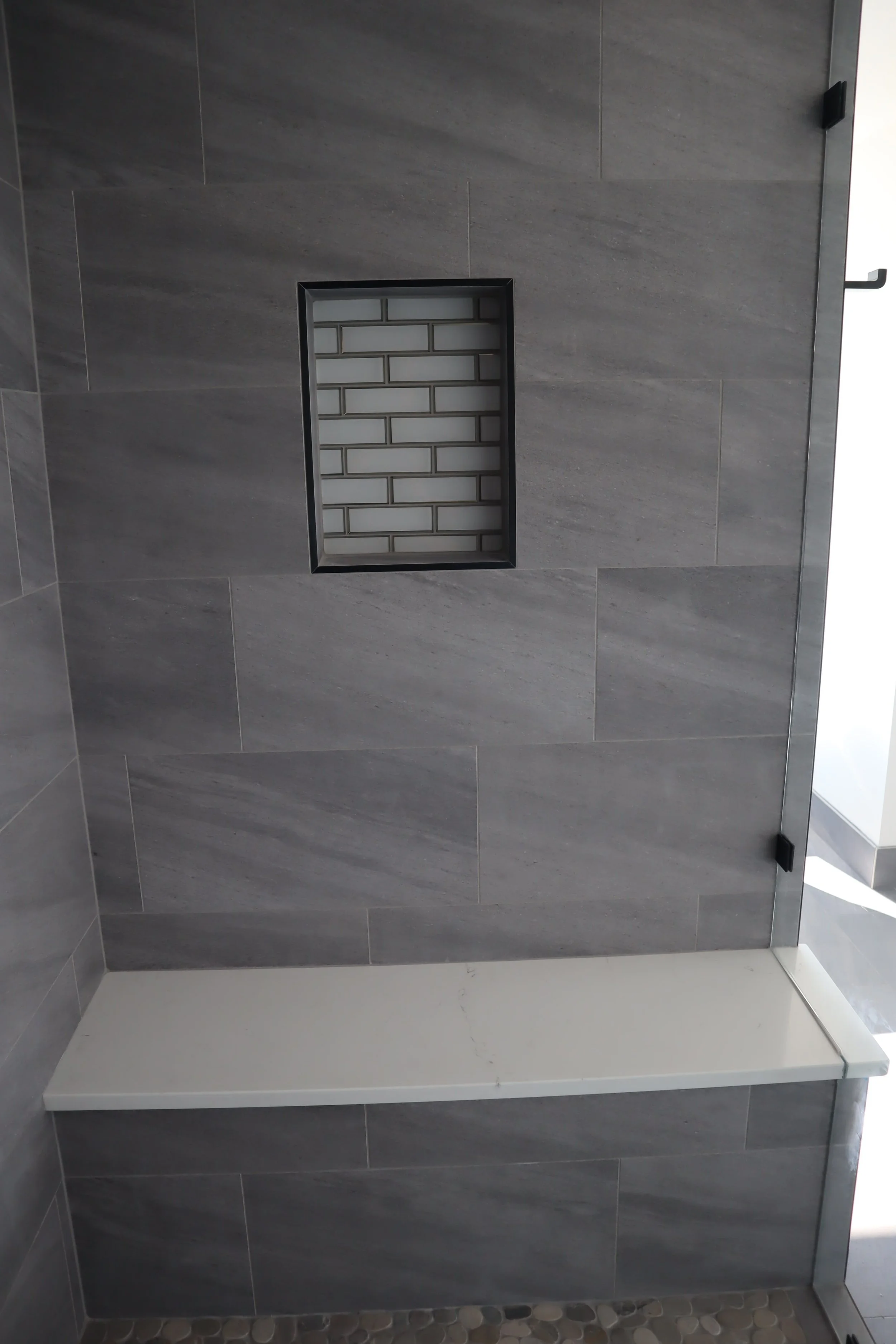 Modern shower with gray tile wall, built-in recessed shelf with white subway tiles, bench seat, and pebble stone floor.
