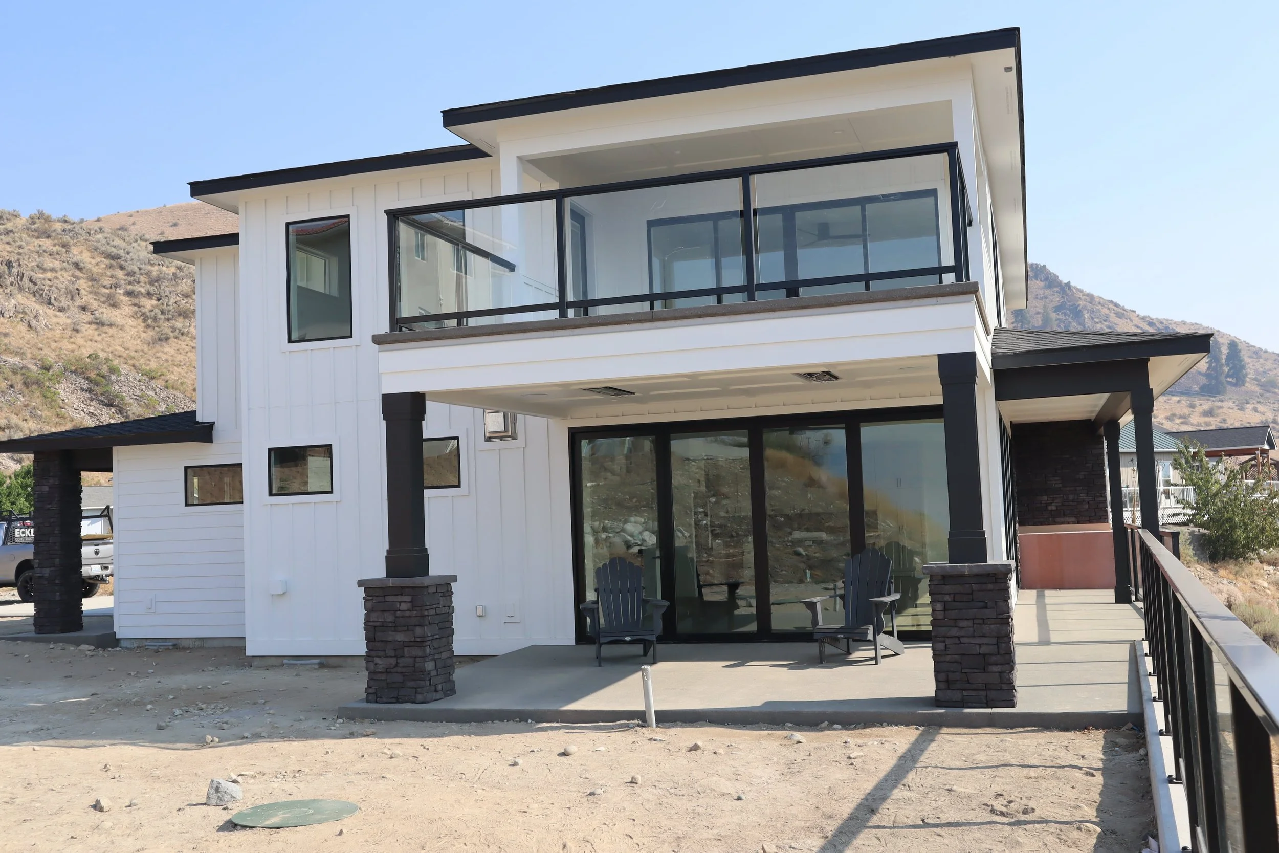 Modern two-story house with a glass balcony, patio with chairs, and stone pillars, set against a rocky hillside.