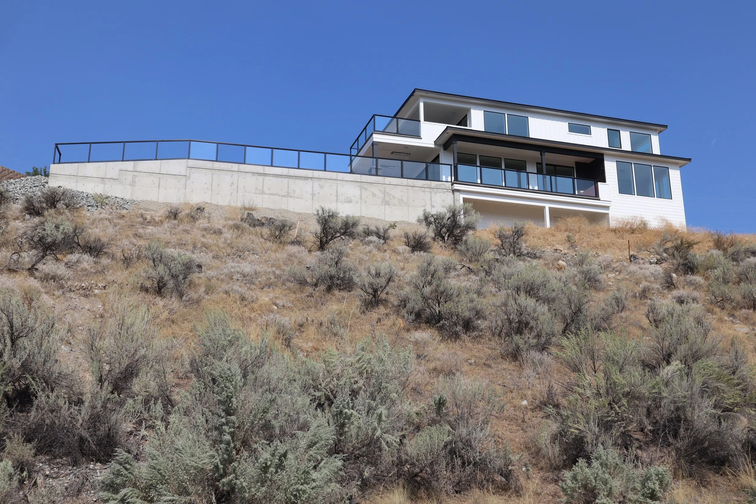 Modern house on a hill with dry vegetation and concrete retaining wall, under a clear blue sky.