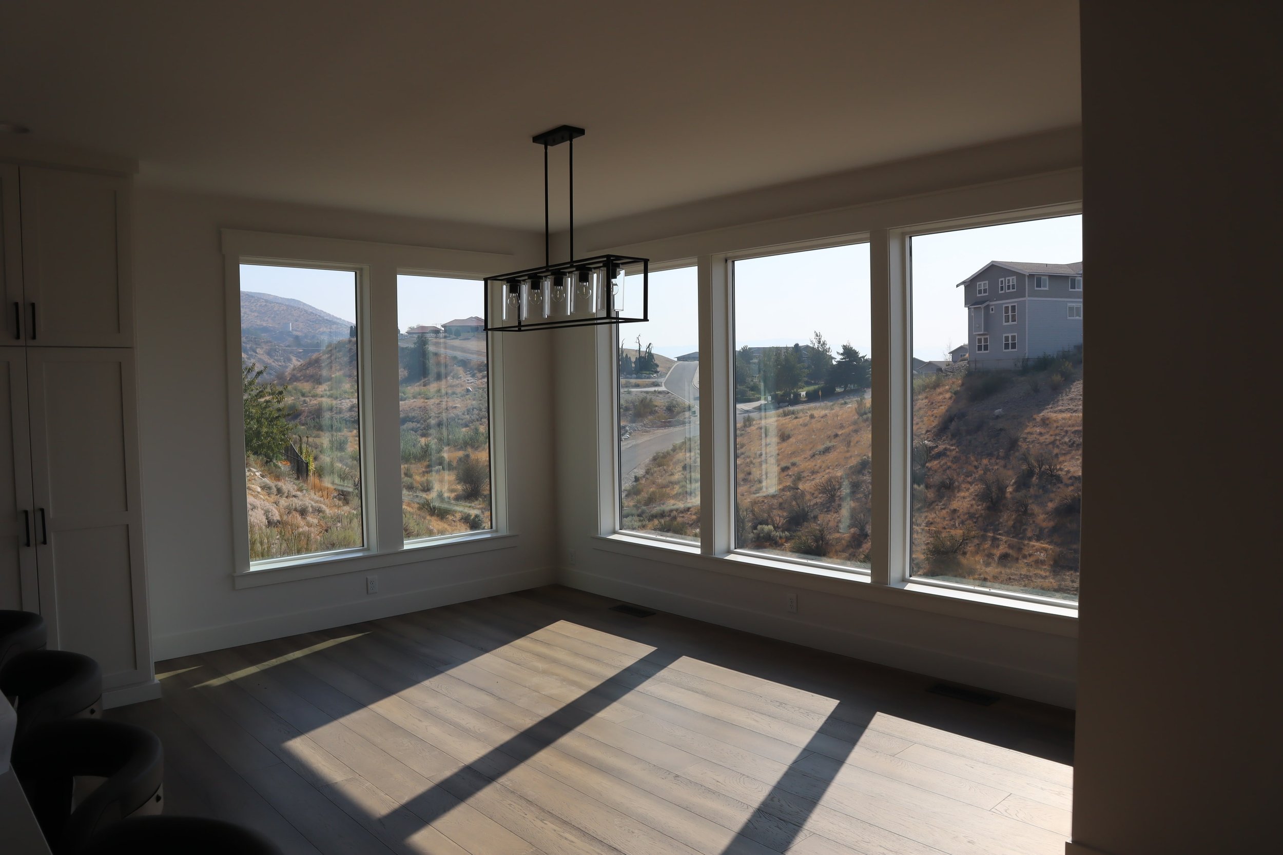 Interior of a modern room with large windows, offering a view of a hilly landscape with a road and a house in the distance. The floor is wooden, and there is a ceiling light fixture.