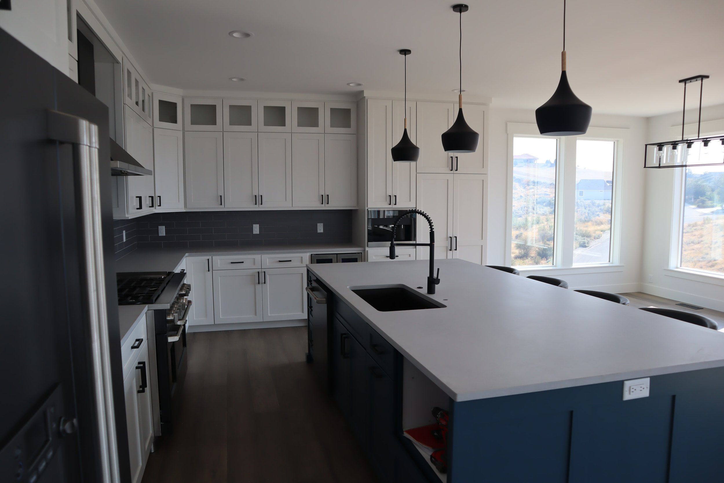Modern kitchen with white cabinets, black countertops, large island, pendant lighting, and a view through large windows.
