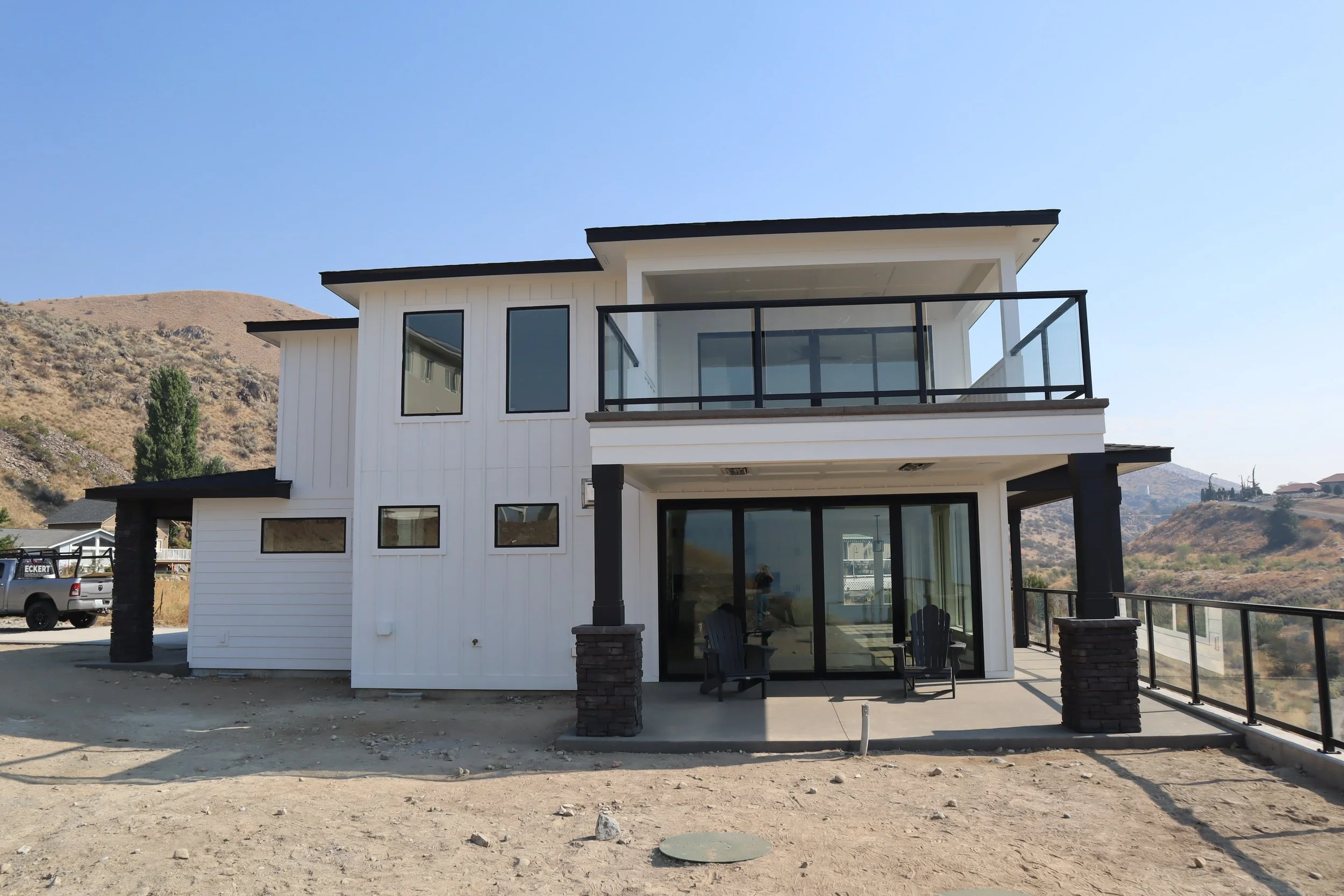 Modern white house with multiple glass windows, balcony, and sliding glass doors, situated in a desert landscape with hills in the background.