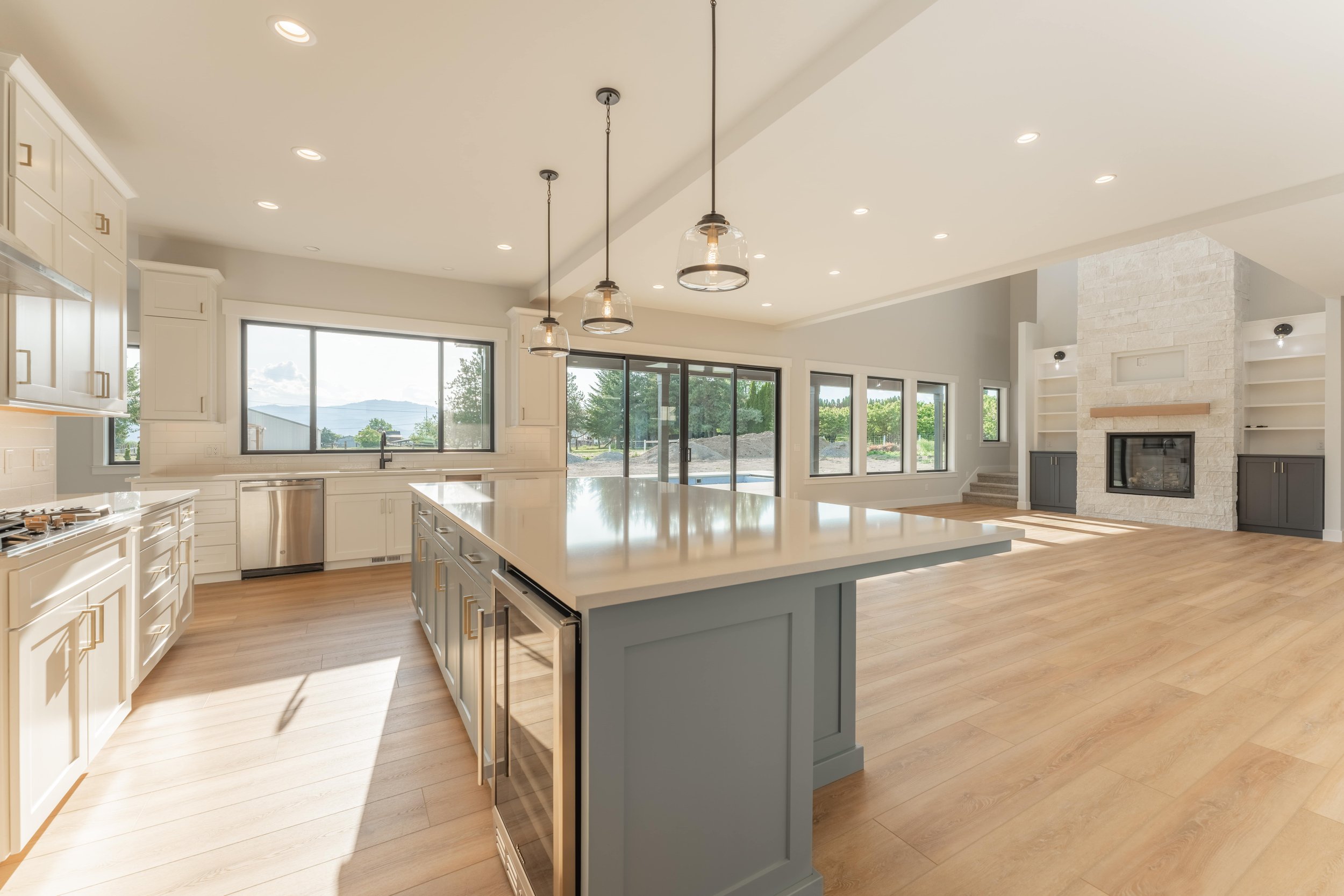Modern kitchen and living area with large island, wood flooring, pendant lights, white cabinets, stainless steel appliances, sliding glass doors, and stone fireplace.