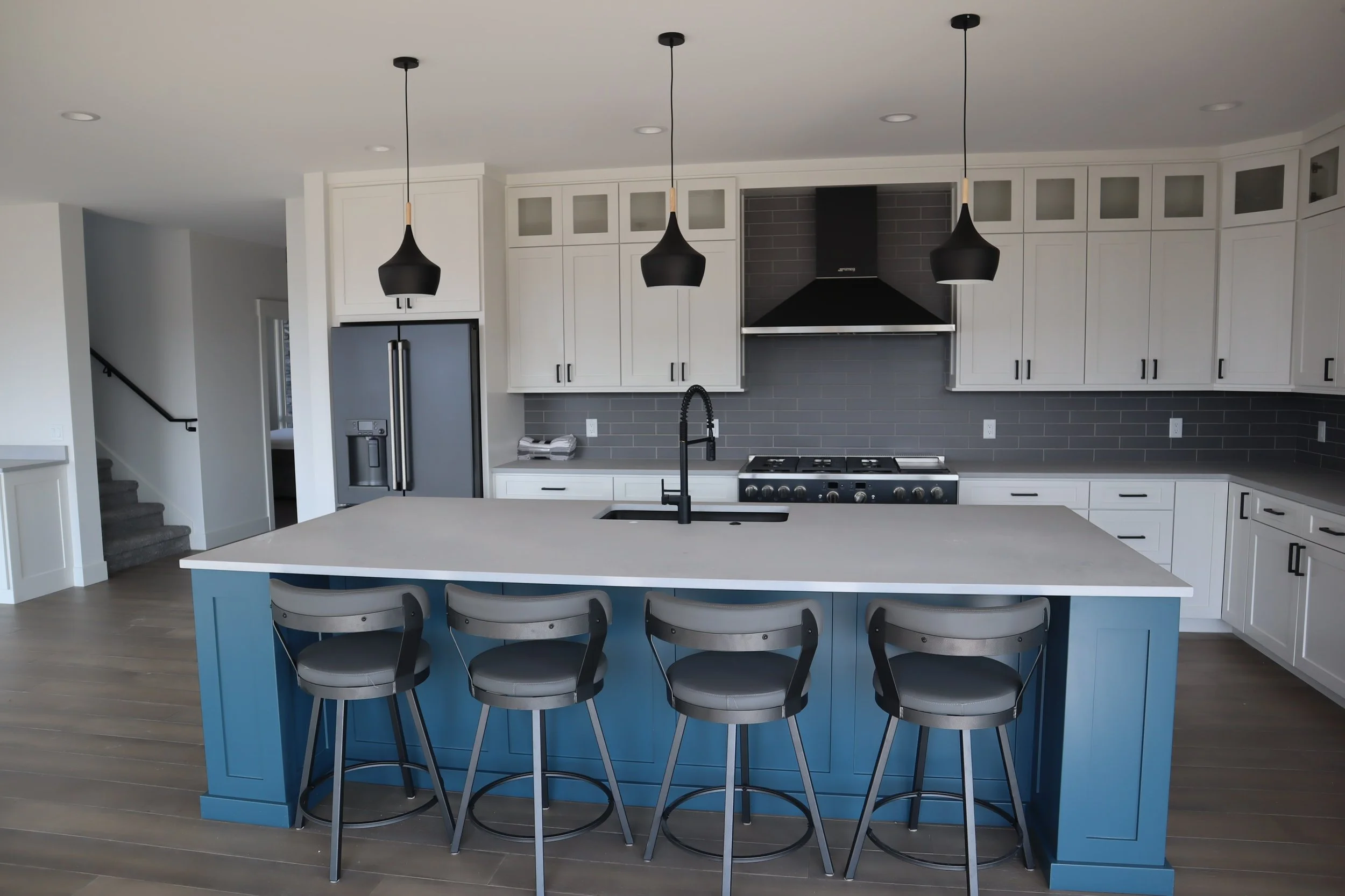 Modern kitchen with white cabinets, blue island, gray tile backsplash, stainless steel refrigerator, stove, and black pendant lights.