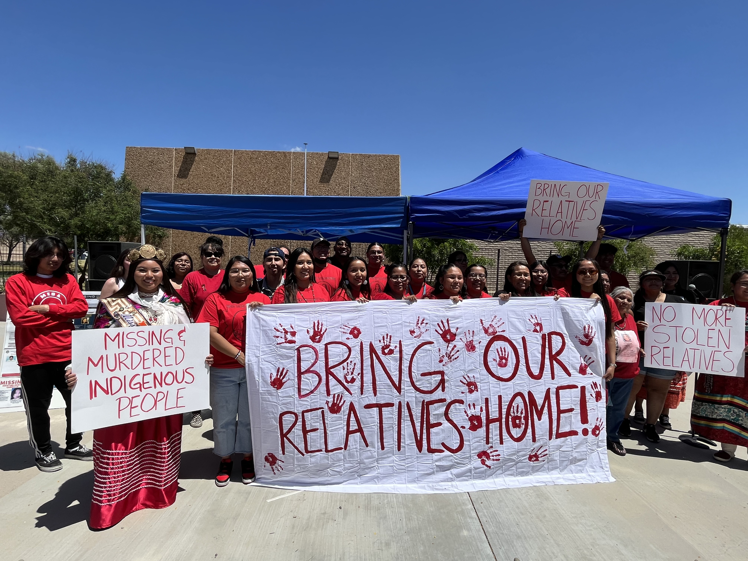 A diverse group of people holding signs and banners at a protest or rally outdoors under a blue sky. The signs contain messages advocating for missing and murdered indigenous people, bringing relatives home, stopping violence against women and girls, and other social justice causes.