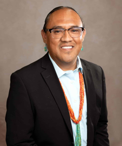 Portrait of a smiling man with glasses, wearing a black blazer, a white shirt, and traditional jewelry, against a neutral background.