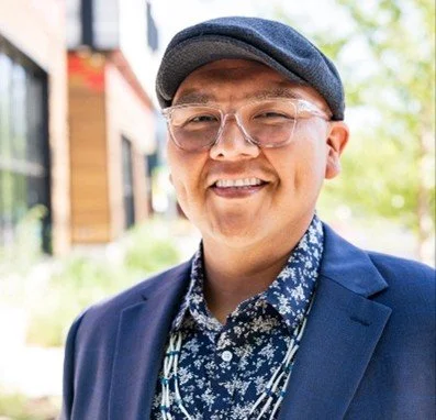 A smiling man wearing glasses, a blue blazer, a patterned shirt, and a flat cap, standing outdoors with trees and a building in the background.