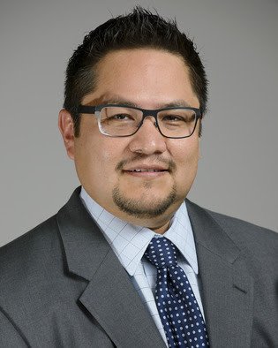 Professional portrait of a man in a suit and tie wearing glasses, smiling against a plain background.