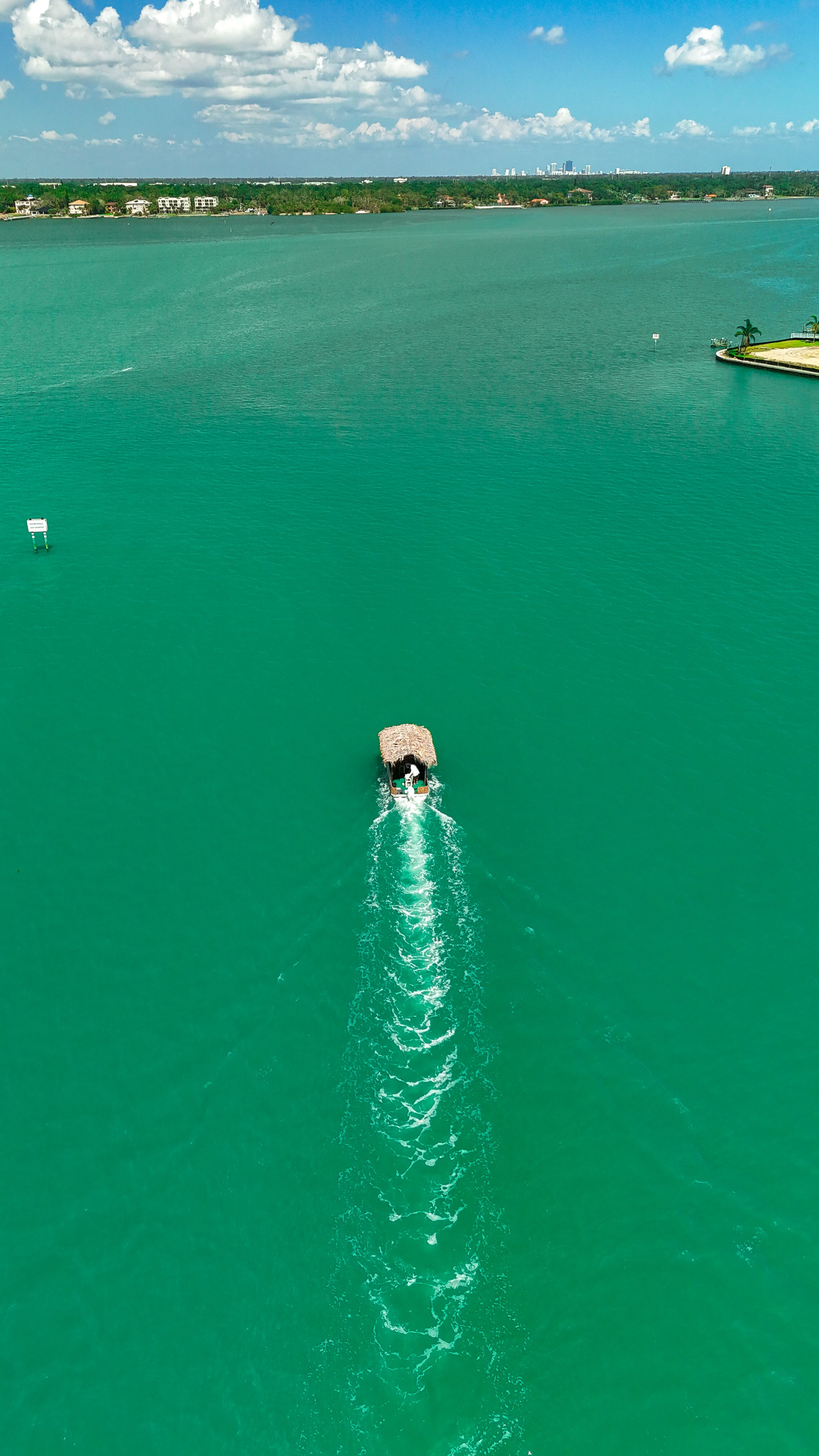A boat with a thatched roof is sailing across a large green lake, leaving a trail in the water. In the background, there are houses along the shoreline and a city skyline with tall buildings under a partly cloudy sky.