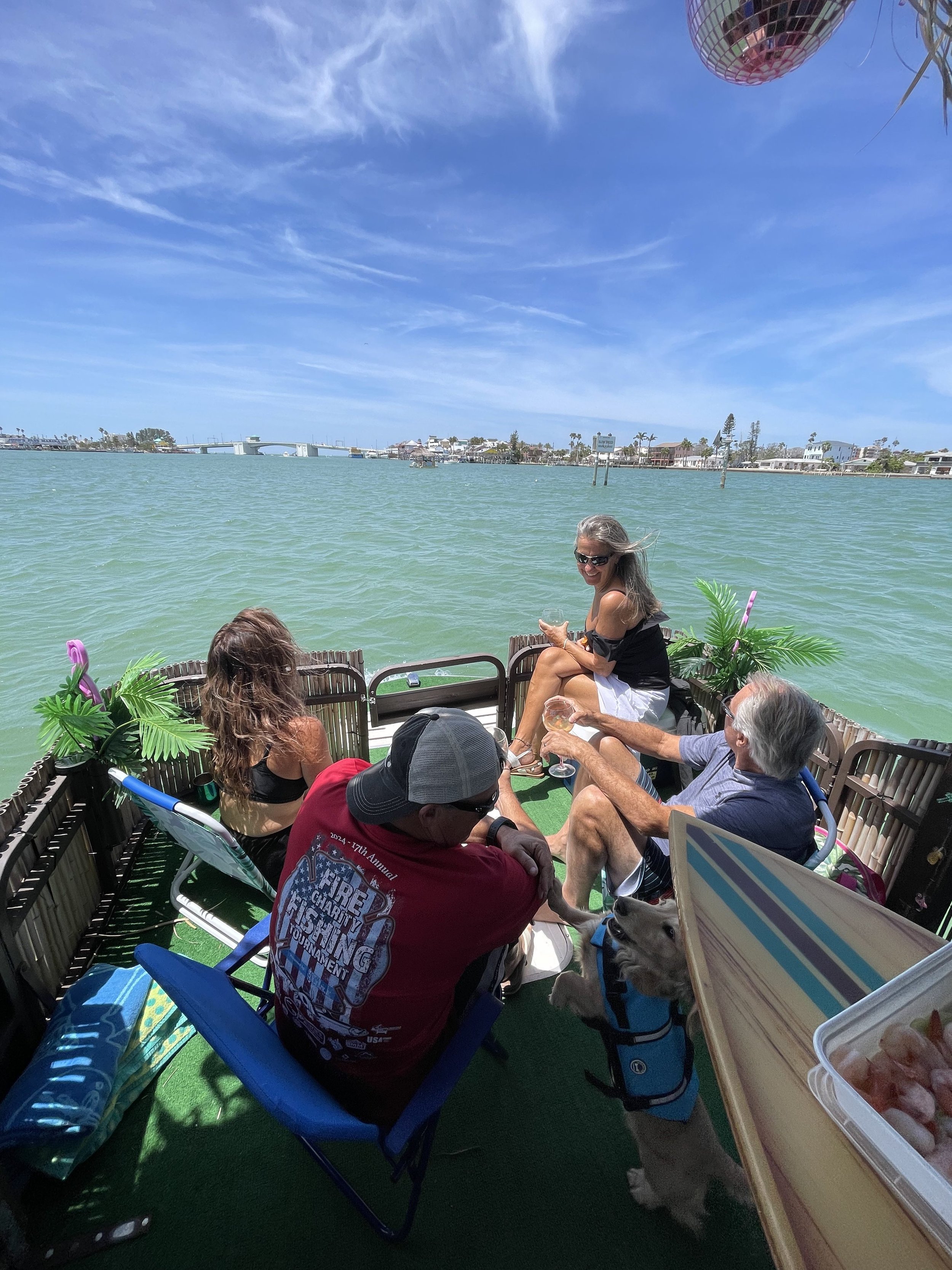 People relaxing on a boat with a dog wearing a life vest, overlooking a scenic view of water and coastal buildings in the background.