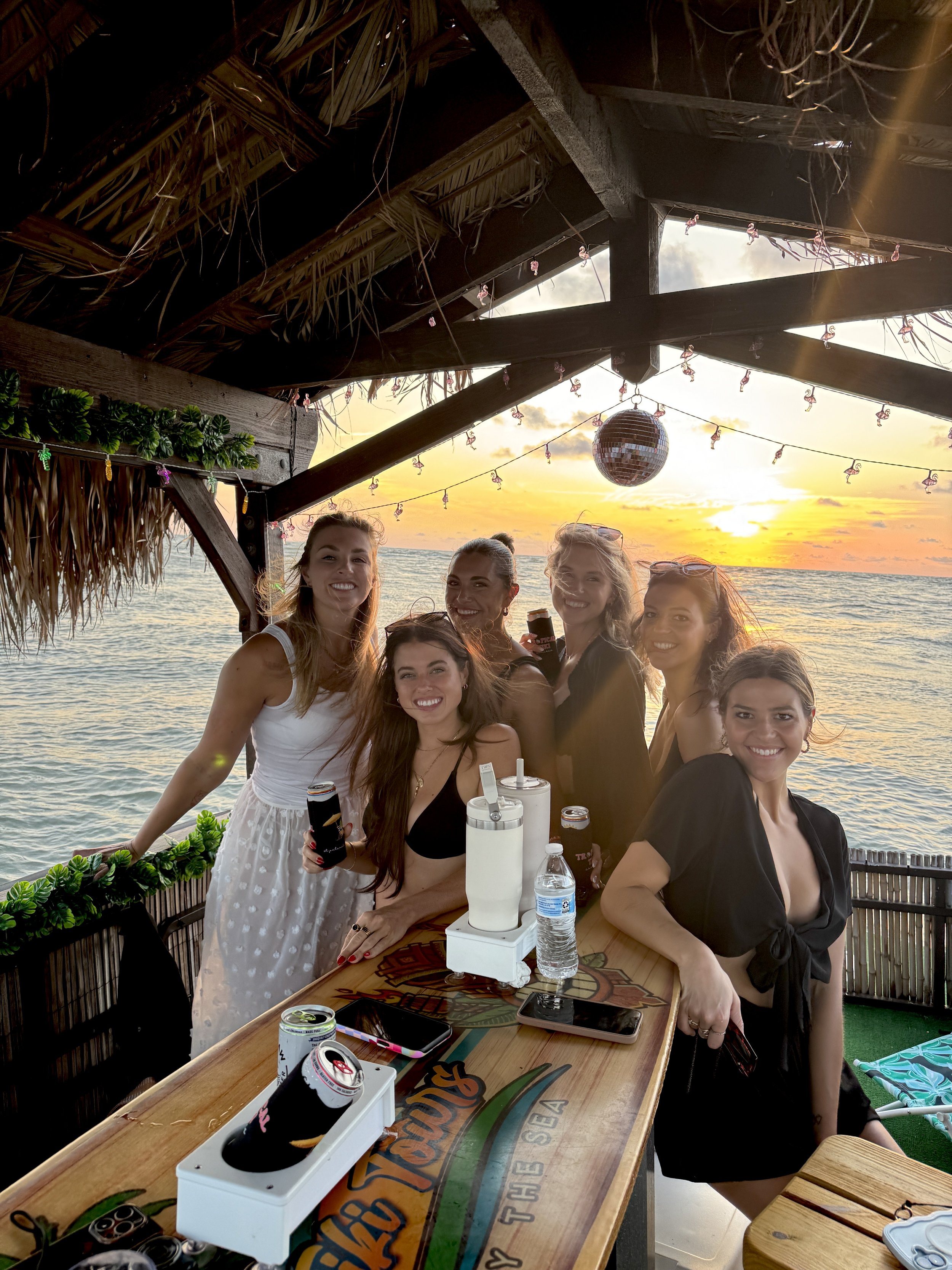 Group of six women celebrating at a beachside bar during sunset, with drinks and electronic devices on the table, under a thatched roof with a disco ball hanging, overlooking the ocean.