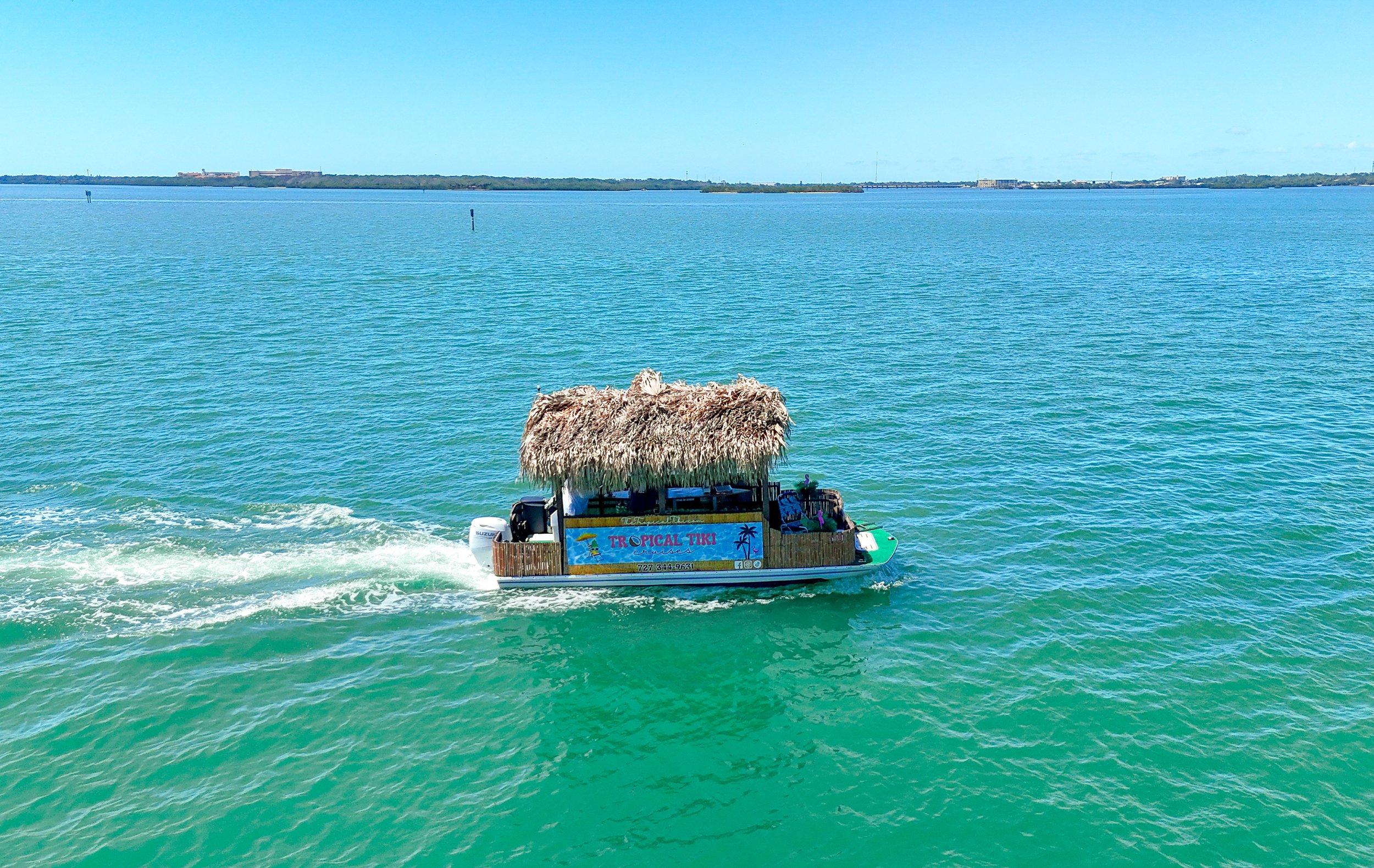 A small boat with a thatched roof bar, labeled 'Tropical Tiki,' is cruising across turquoise water on a sunny day, with land visible in the background.