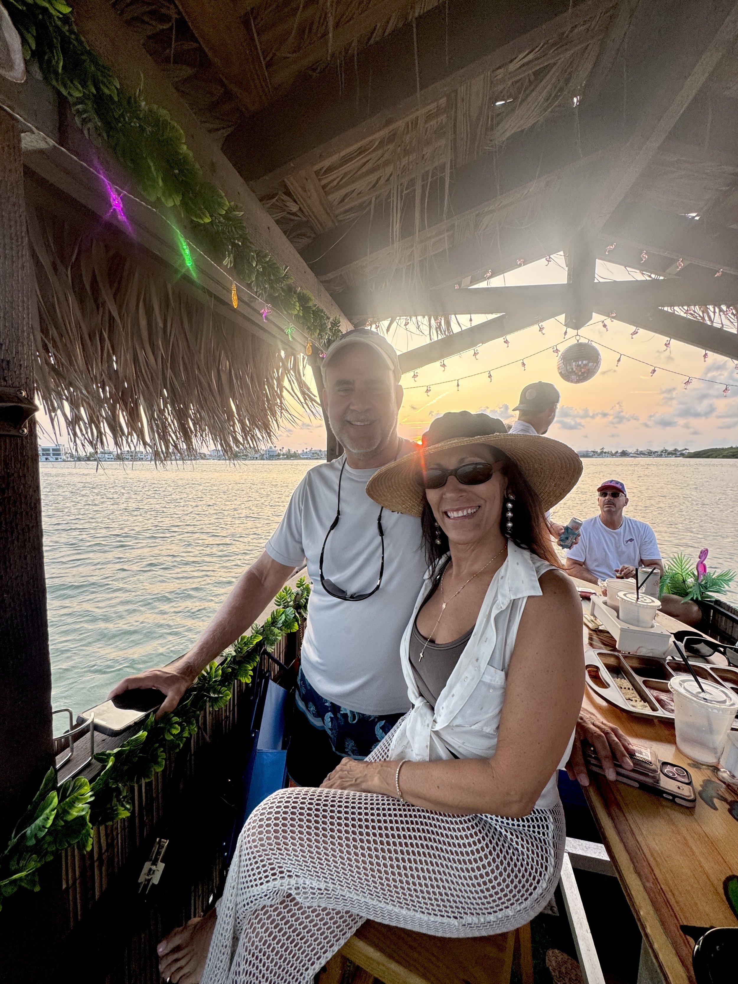 A man and a woman are smiling on a boat at sunset, with water and a scenic sky in the background. The woman is wearing a large straw hat, sunglasses, and a white outfit, while the man is in a white shirt and Shorts. There are drinks and snacks on the