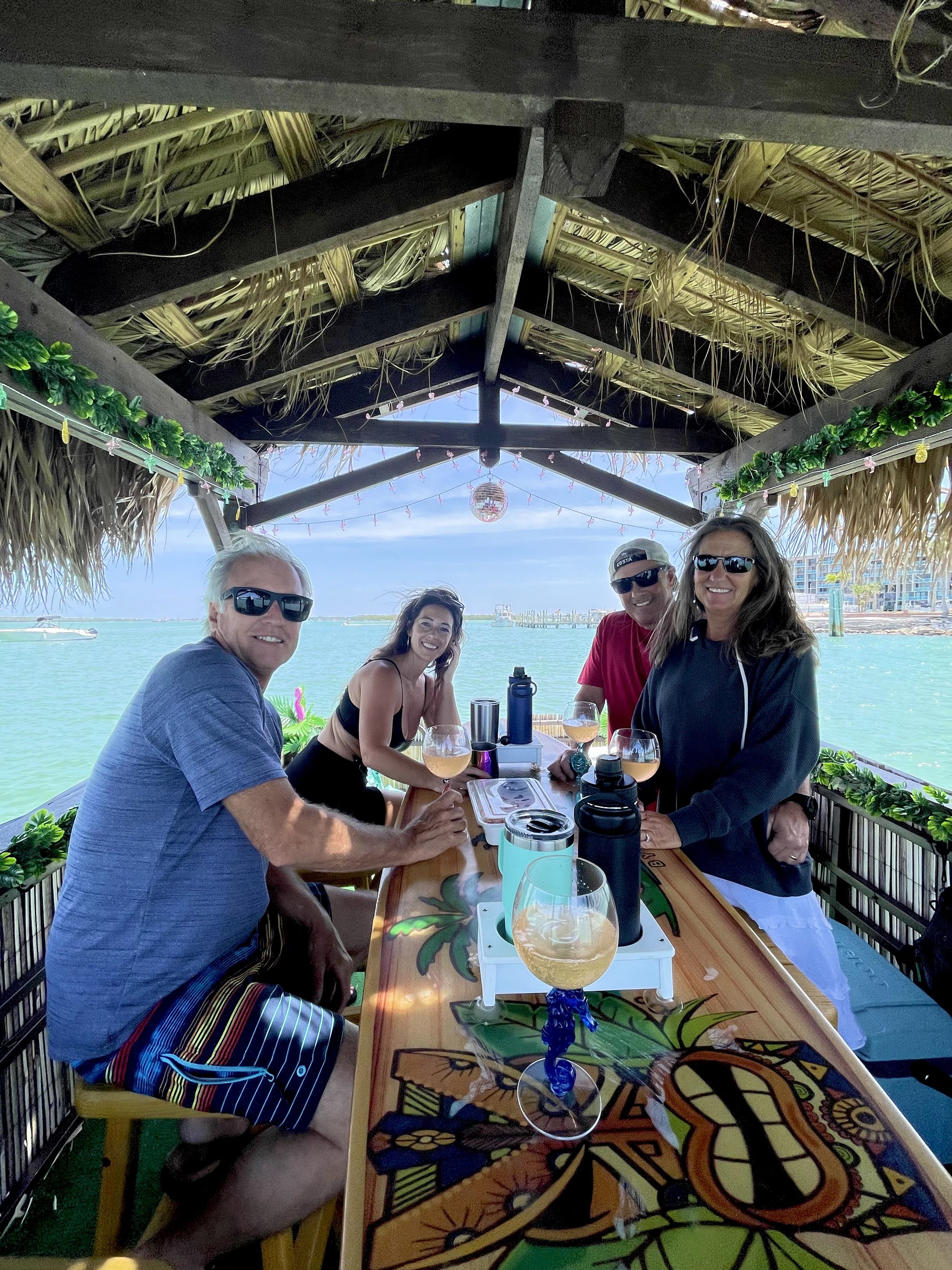 Group of four people sitting at a tiki bar on the water, enjoying drinks, under a thatched roof with tropical decorations.