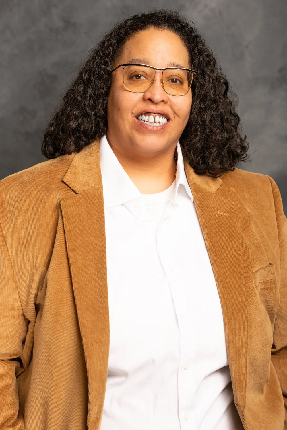 Chauntyll Allen wearing glasses, white shirt, and tan blazer, smiling against a gray background.