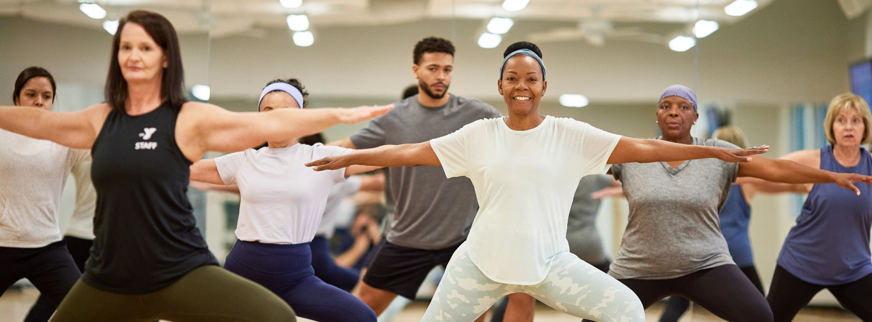 A diverse group of people participating in a fitness or yoga class, led by an instructor at the front, in a well-lit indoor studio.