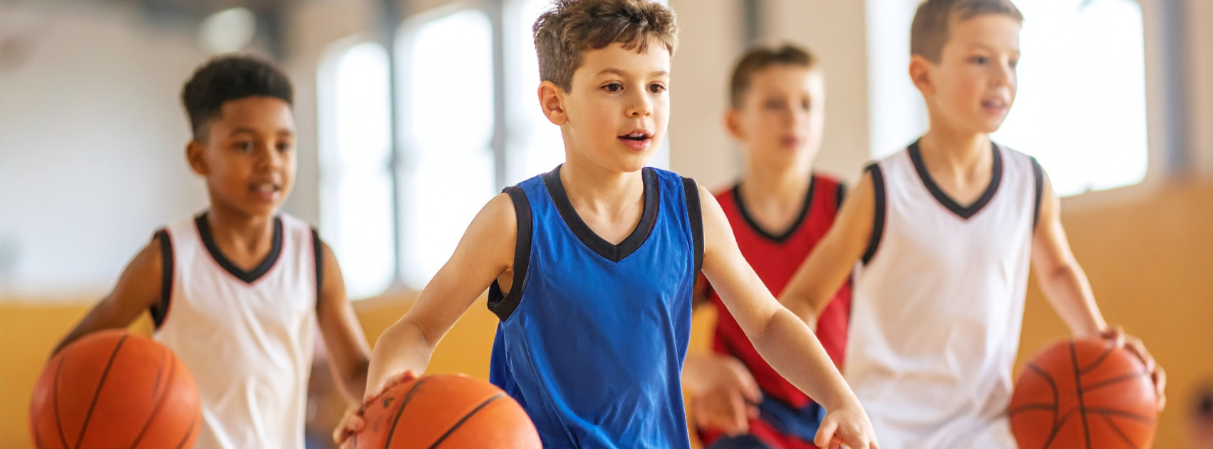 Four young boys playing basketball in an indoor gym, wearing sports uniforms.