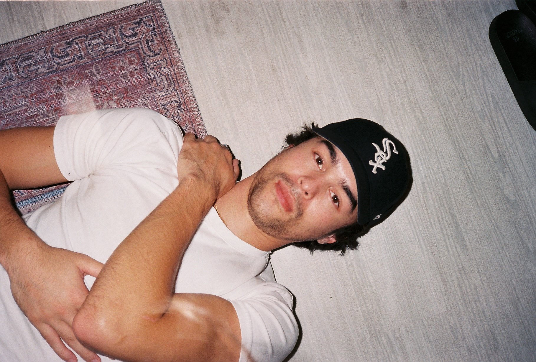 A young man lying on a patterned rug on the floor against a light-colored wall, wearing a black baseball cap and a white t-shirt, with his left hand across his chest and his right hand touching his chin.