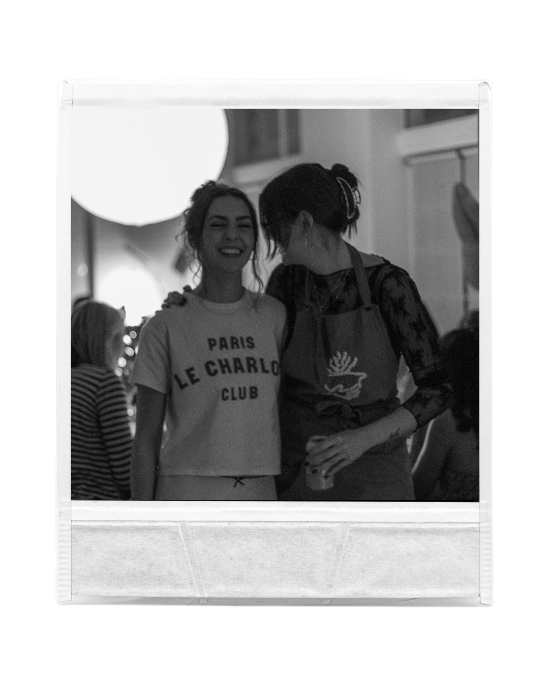 Black and white photo of two women smiling and sharing a moment at a social gathering, one wearing a T-shirt with French words, and the other in a lacy top with a scarf, surrounded by other people.
