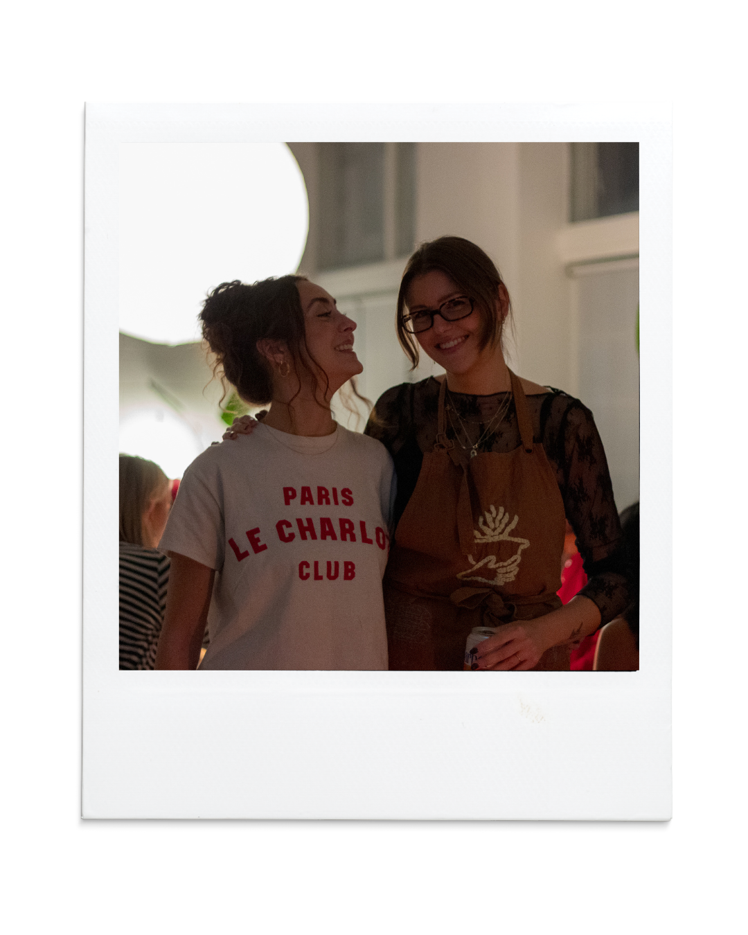 Two women smiling and standing close together at an indoor gathering, one wearing a white t-shirt with red text and the other wearing glasses and a brown apron.