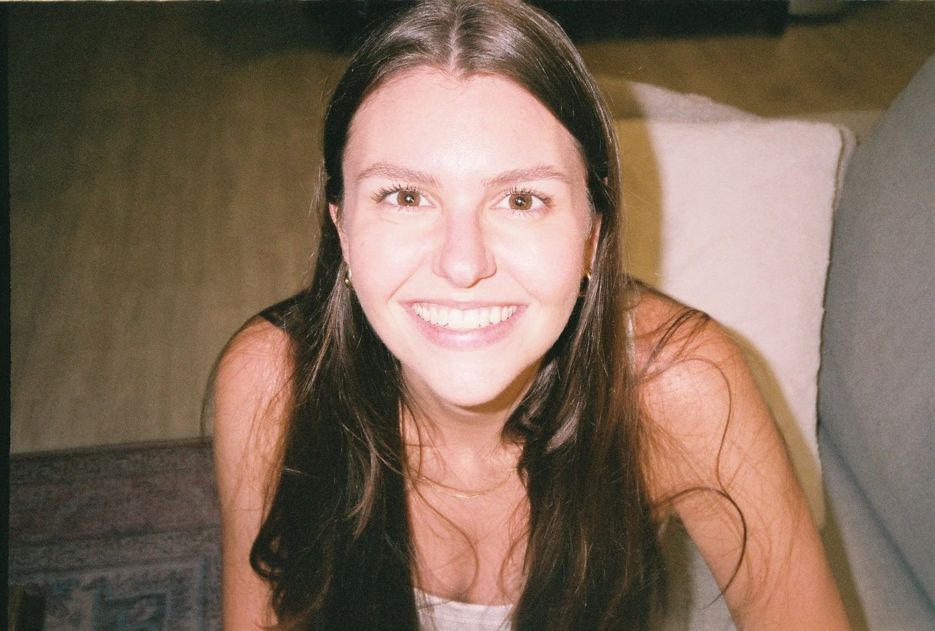 Close-up of a young woman with long brown hair, brown eyes, and a big smile, wearing earrings and a delicate necklace, sitting on a couch with a pillow in the background.