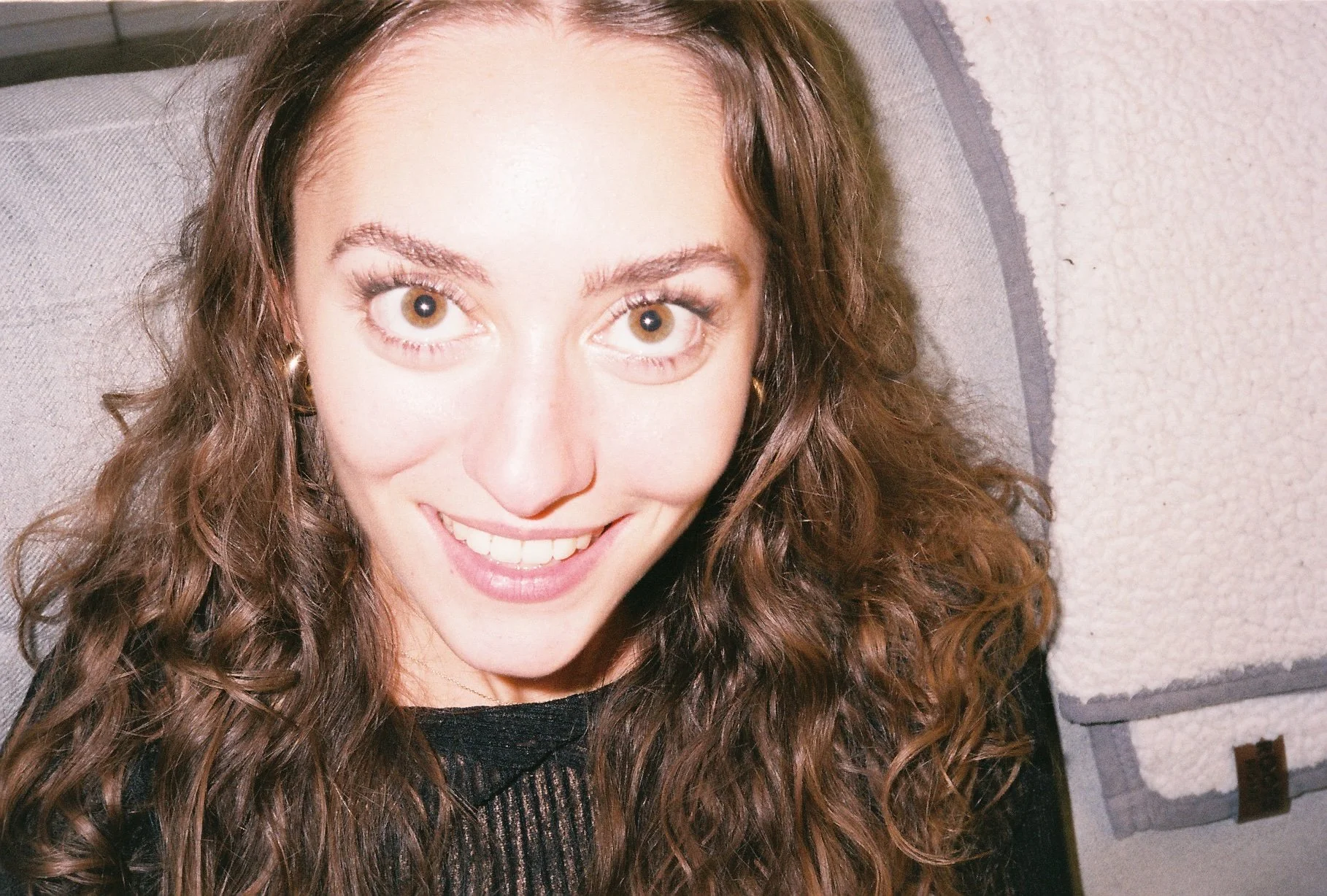 Close-up of a woman with brown curly hair smiling, wearing a black top, in front of a textured wall and a gray cushion.