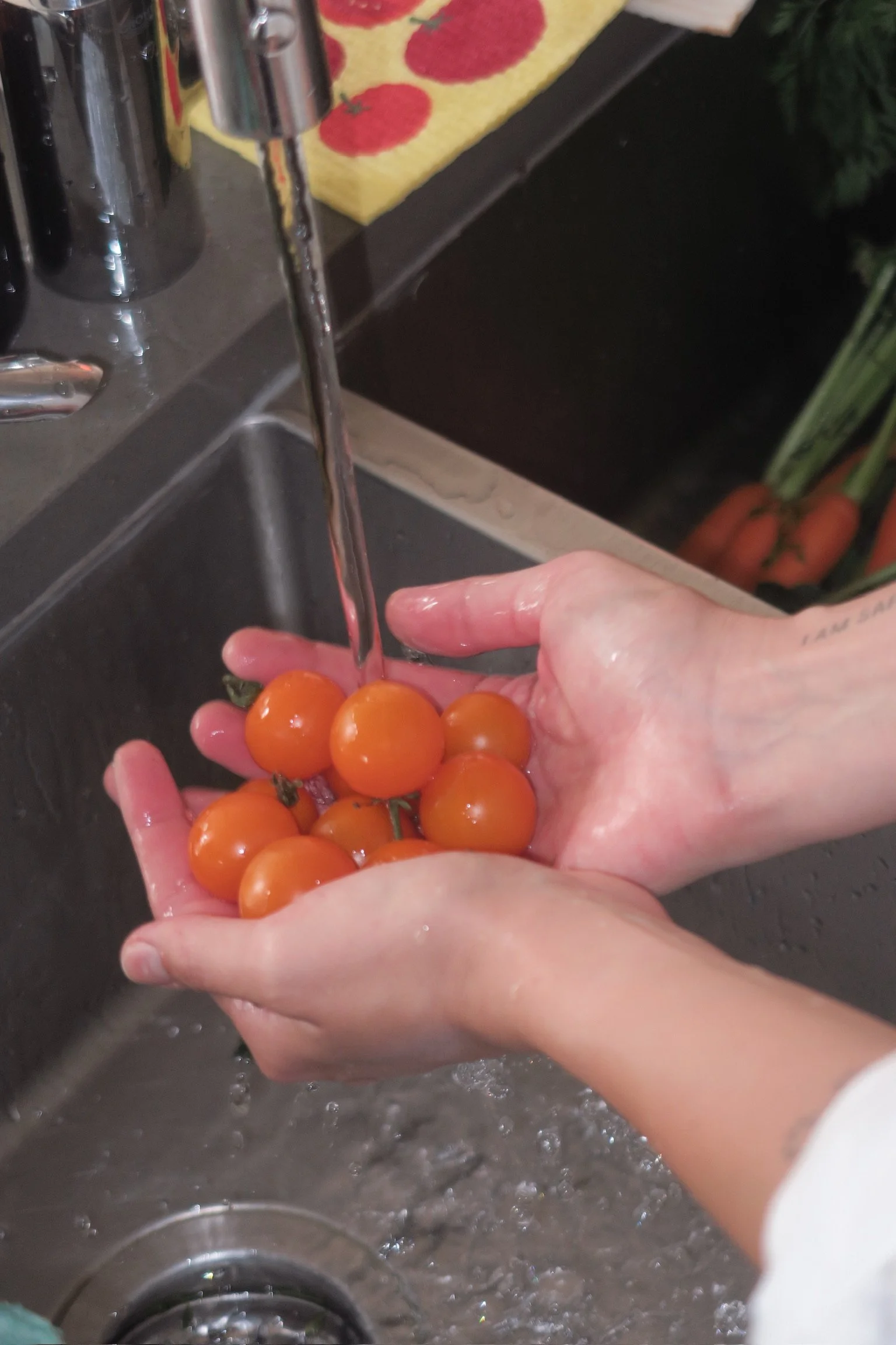 A person washing cherry tomatoes under running water in a kitchen sink.