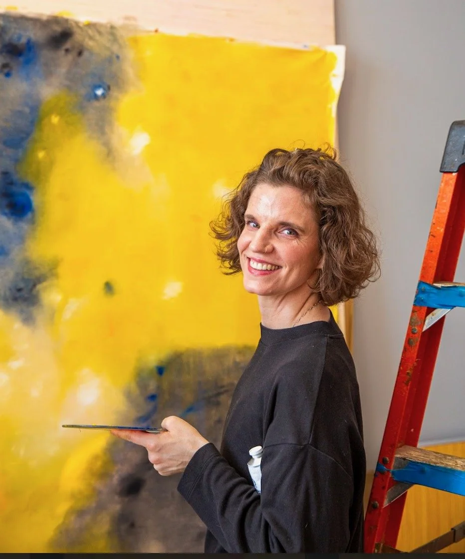 Adriane R Wiltse turns to you, smiling, wearing all black, holding a paint pallet and brush with a golden yellow painting behind.