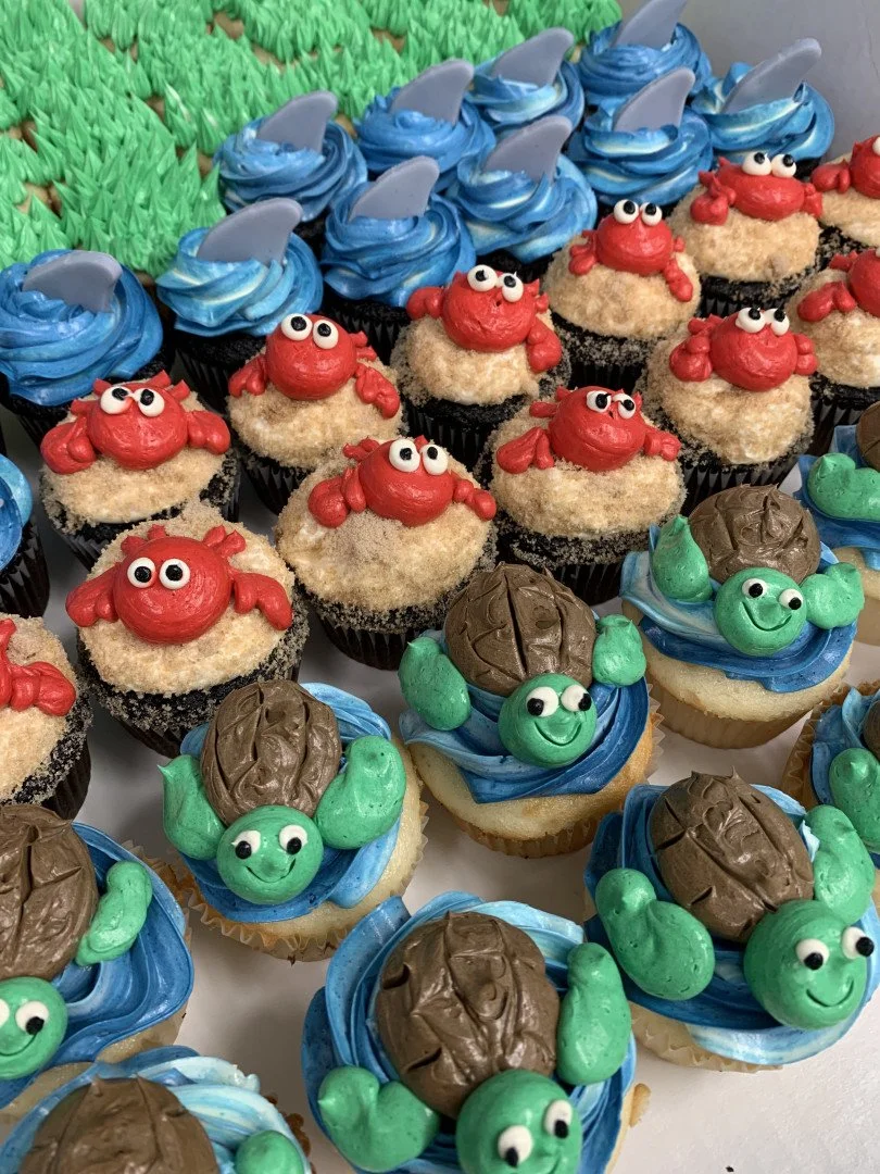 Assorted cupcakes decorated with ocean-themed designs, including red crabs with white eyes, green turtles with brown shells and smiling faces, and blue swirled frosting.