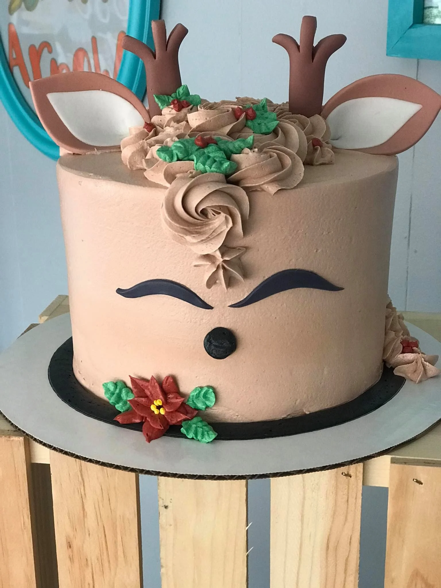 A cake decorated to resemble a reindeer, featuring antlers, ears, closed eyes, a red poinsettia flower at the base, and holly leaves with red berries on top.